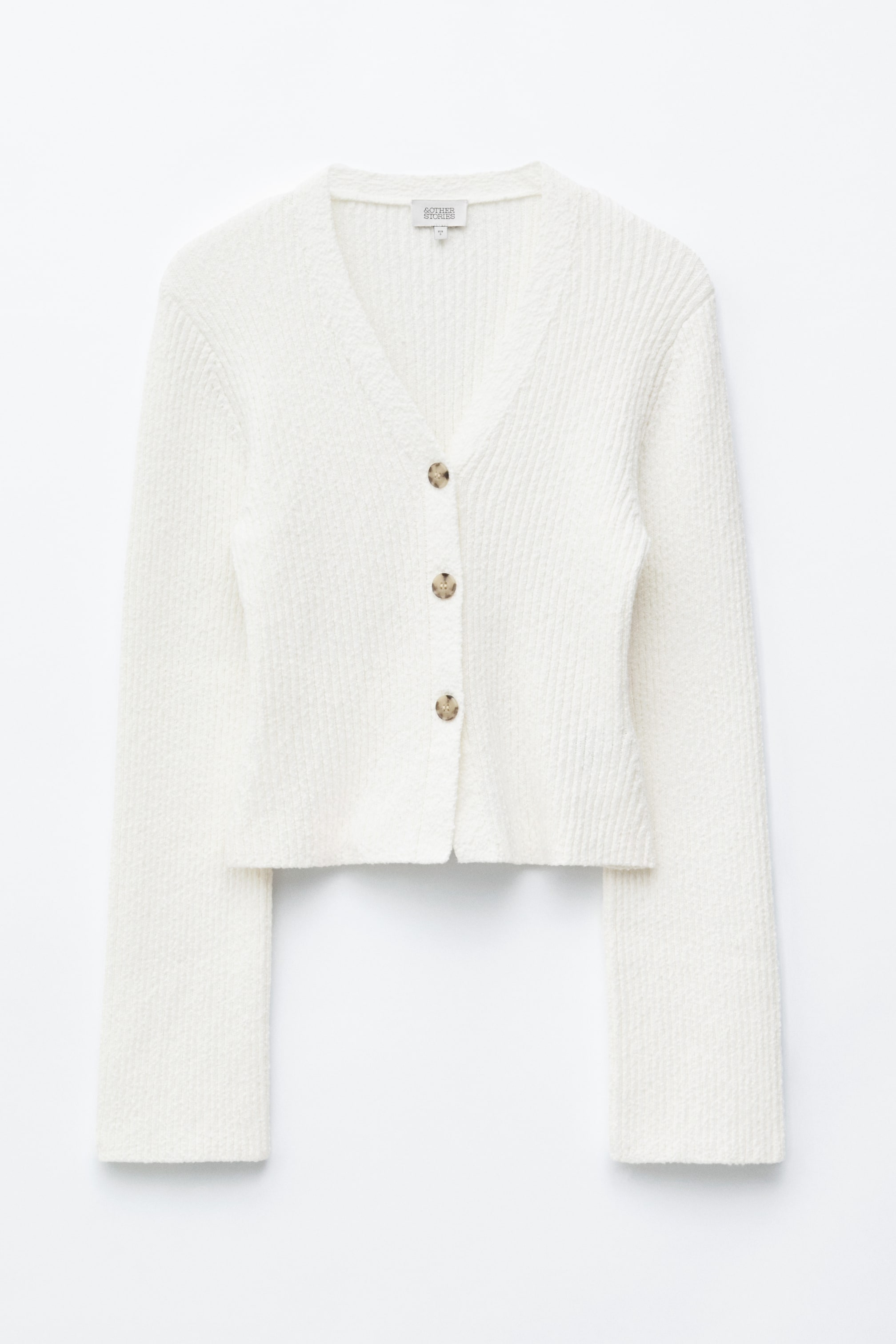 Se større billede: En hvid cardigan viser sin front, med en V-halsudskæring og en ribbet knappet stolpe med tre lysebrune knapper. Den fremviser et tekstureret, ribbet stof og præsenterer en let cropped længde med lange ærmer.