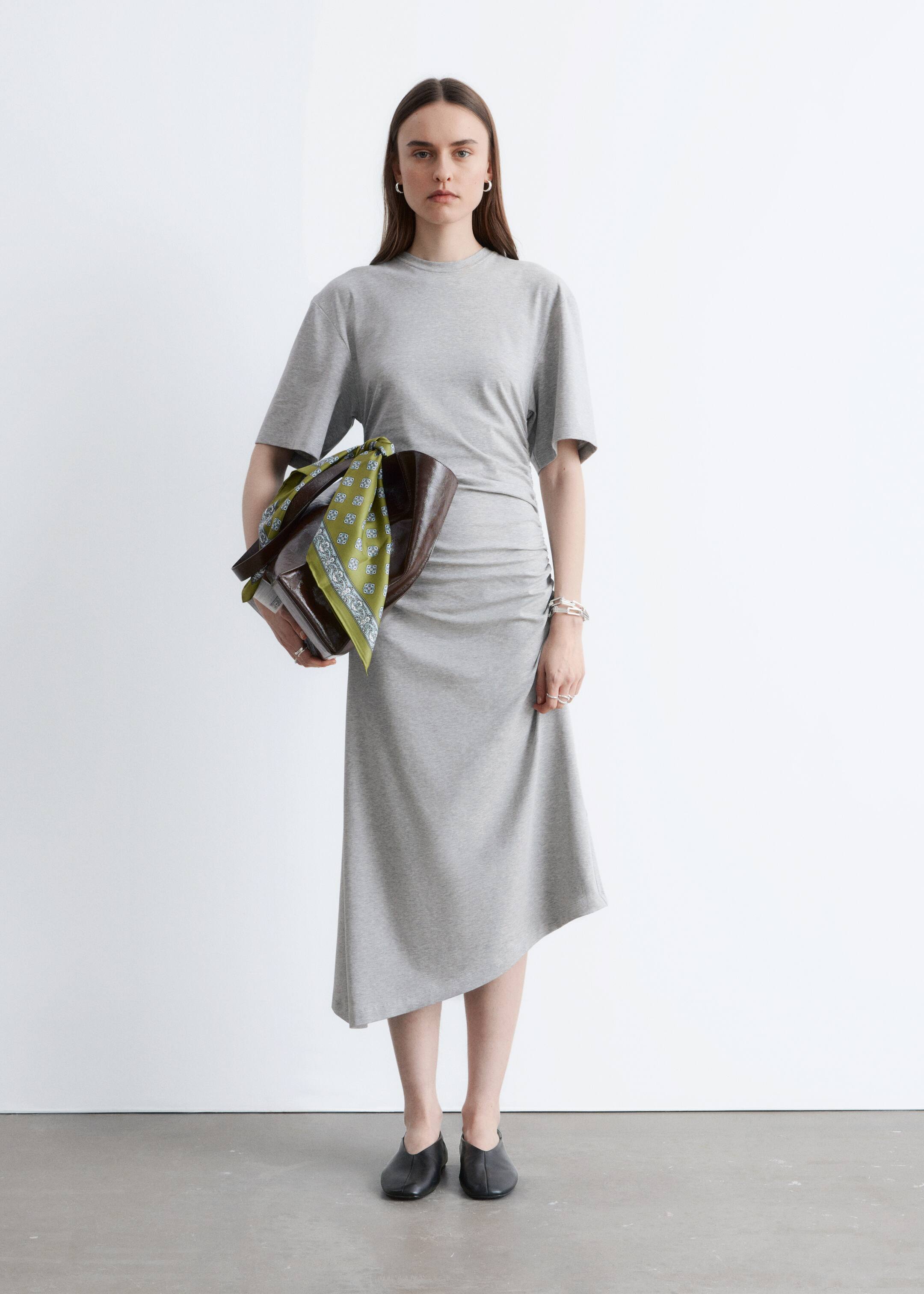 Visualizza immagine più grande: Un abito midi in jersey grigio chiaro presenta uno scollo tondo, maniche corte e arricciature sul lato destro della vita che si trasformano in un orlo asimmetrico.