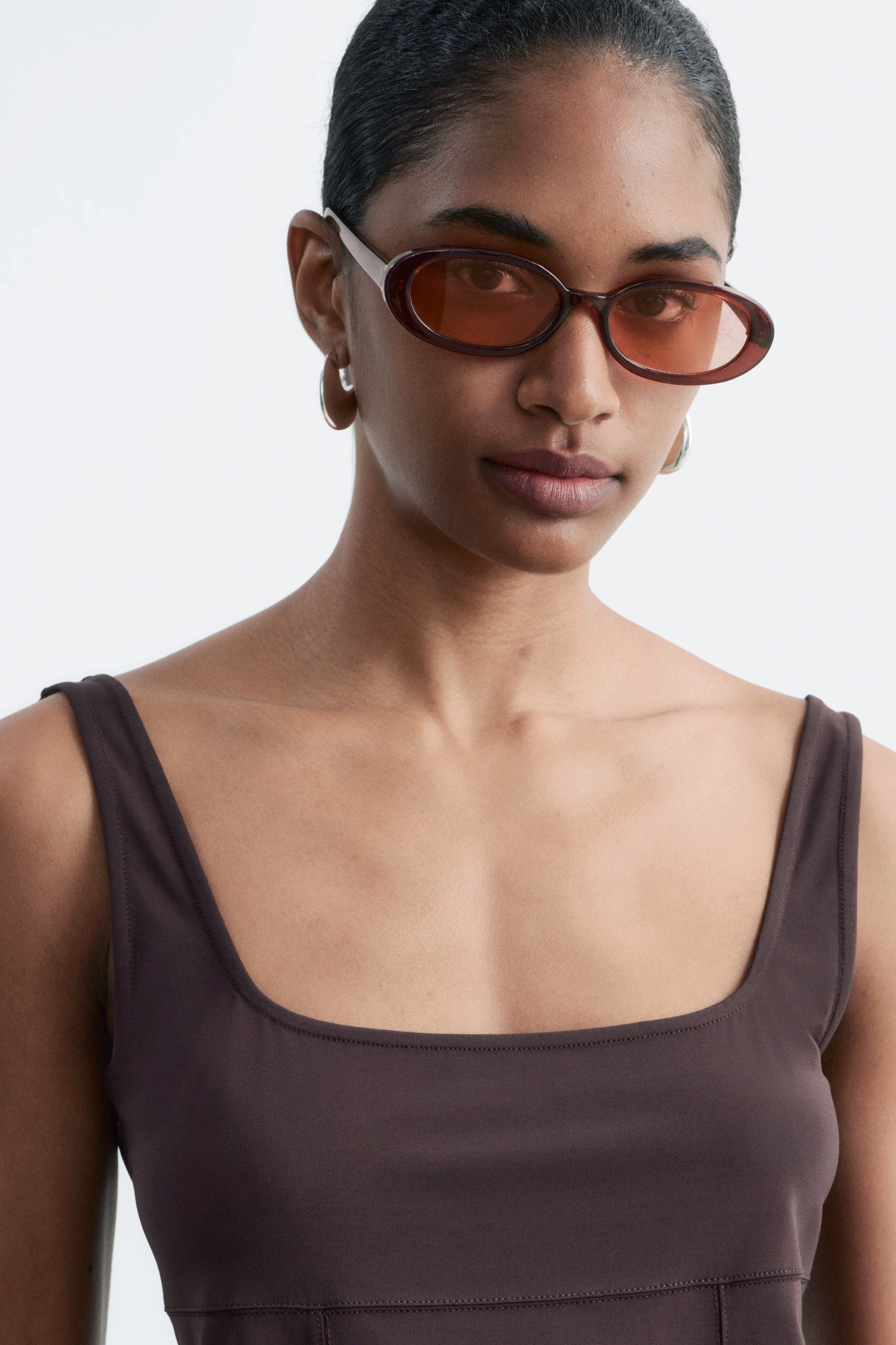 הצגת תמונה גדולה יותר: משקפי שמש אובאליים בצבע חום כהה ומבריק עם עדשות כהות נלבשים על ידי הדוגמנית, שלובשת גם גופיית טנק בצבע חום כהה עם מחשוף מרובע, רצועות כתף רחבות ותפרים גלויים עם קווי חיתוך.
