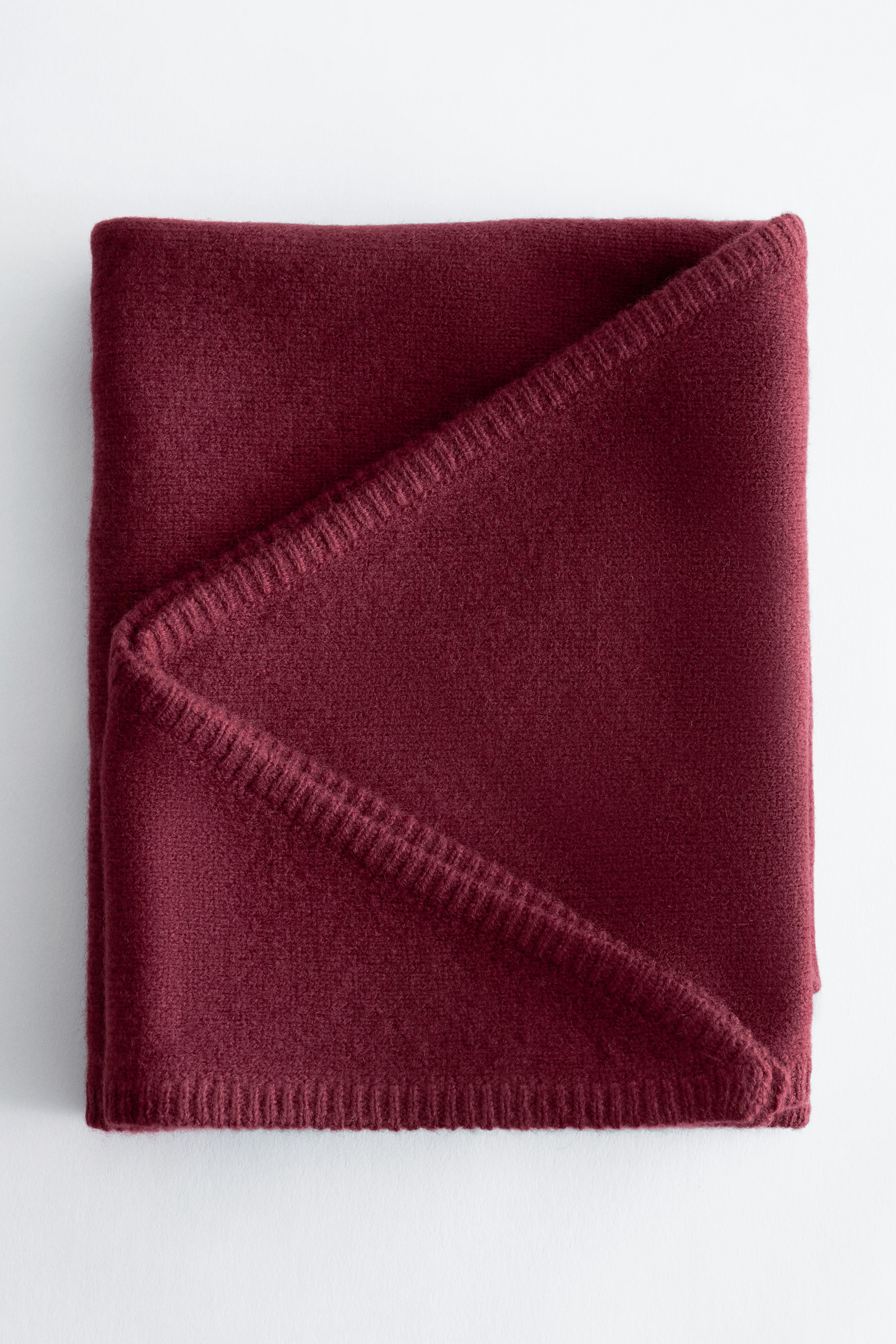 Agrandir l'image: Une couverture rouge foncé est soigneusement pliée, mettant en valeur sa surface douce et texturée. Un coin est replié en diagonale, révélant un bord tricoté visible.