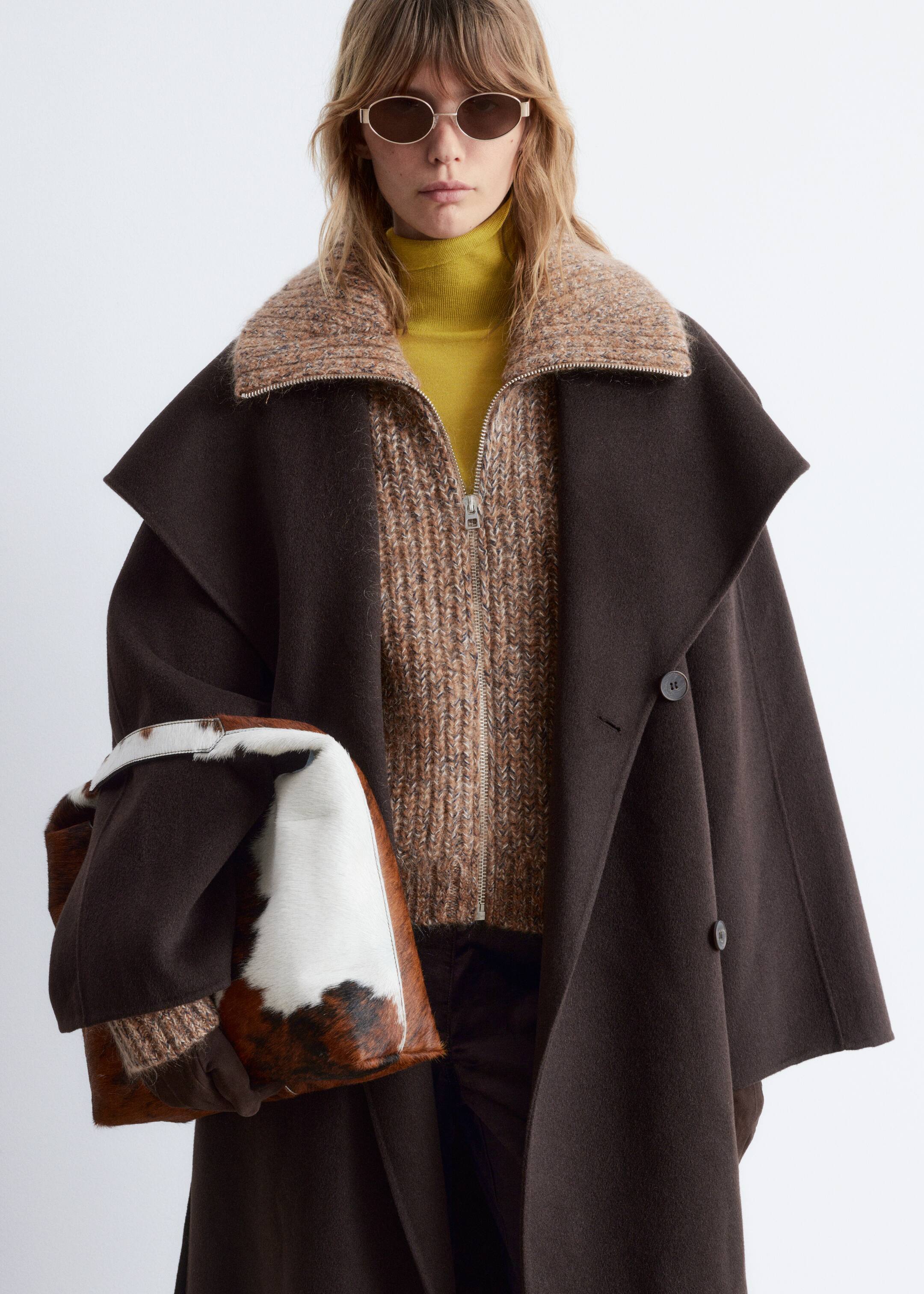Agrandir l'image: Un manteau marron foncé, légèrement orienté vers l'avant, présente une silhouette large et drapée avec des épaules structurées et une fermeture croisée à double boutonnage avec deux boutons visibles. Il est superposé à une veste zippée en grosse maille beige et marron avec un col côtelé et un col roulé jaune, avec une pochette à motif peau de vache et des lunettes de soleil ovales.