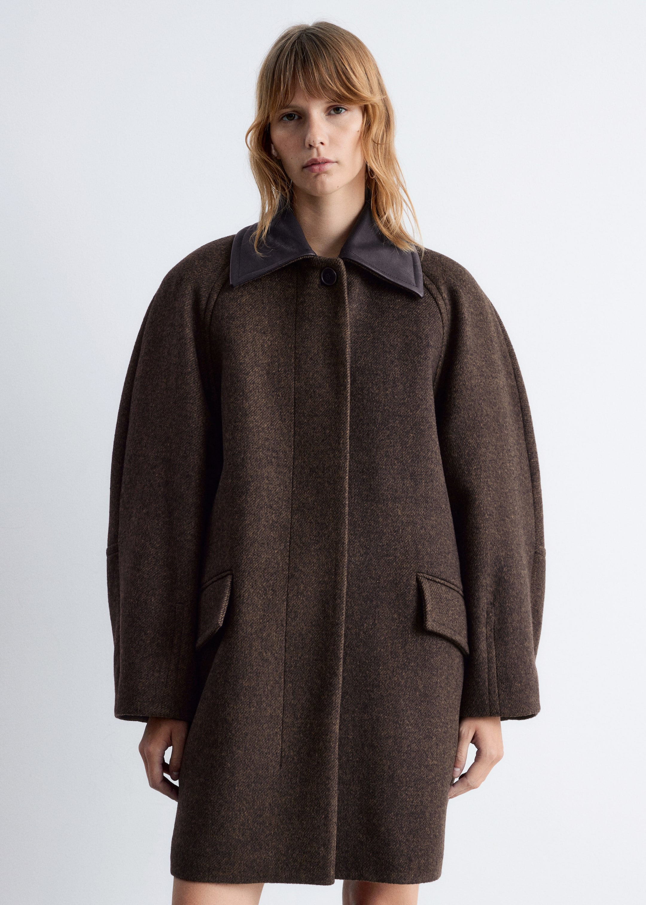 View larger image: A dark brown, textured coat is being displayed from the front, featuring a contrasting dark brown rounded collar and a concealed front button placket. It is designed with voluminous raglan sleeves tapering at the cuffs, and two flap pockets are visible on the front, with the coat falling to an above-knee length.