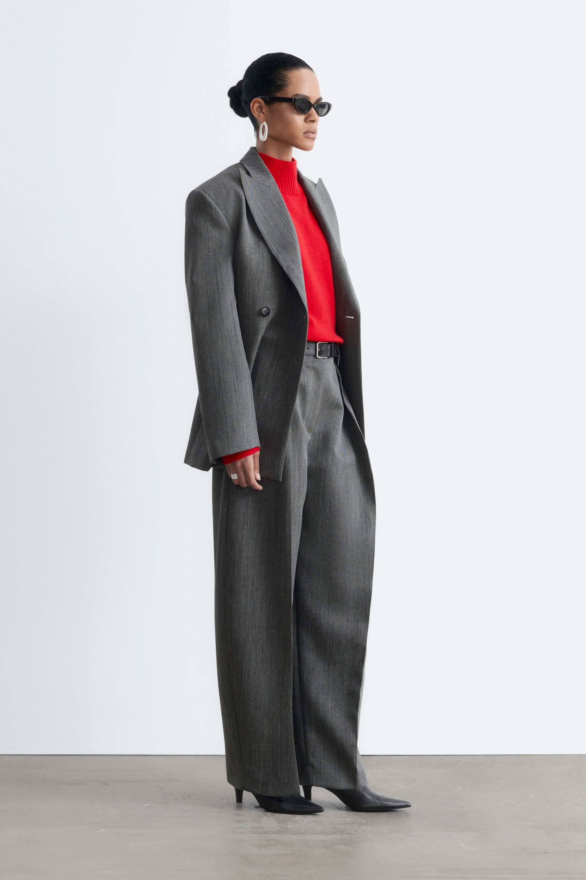 Agrandir l'image: Le mannequin porte un pantalon en laine gris à silhouette barrel leg et avec des plis marqués. Ce pantalon taille haute est associé à une ceinture en cuir noir, à un pull col roulé rouge vif et à un blazer gris assorti.