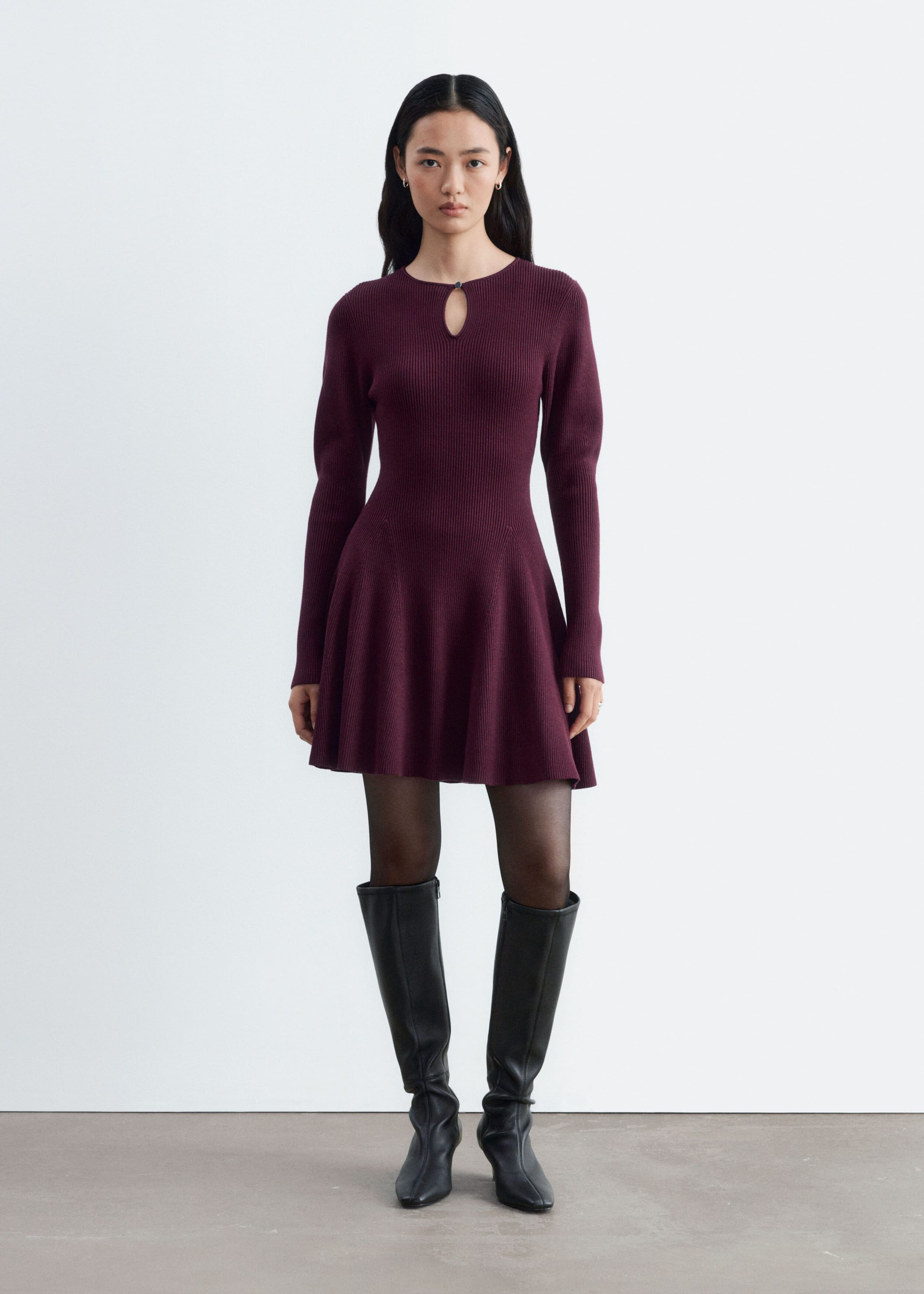 A ribbed knit mini dress in deep plum is being worn by the model, featuring a keyhole neckline with a metal button, long sleeves, and a flared, paneled skirt. The dress is styled with sheer black tights and knee-high black leather boots.