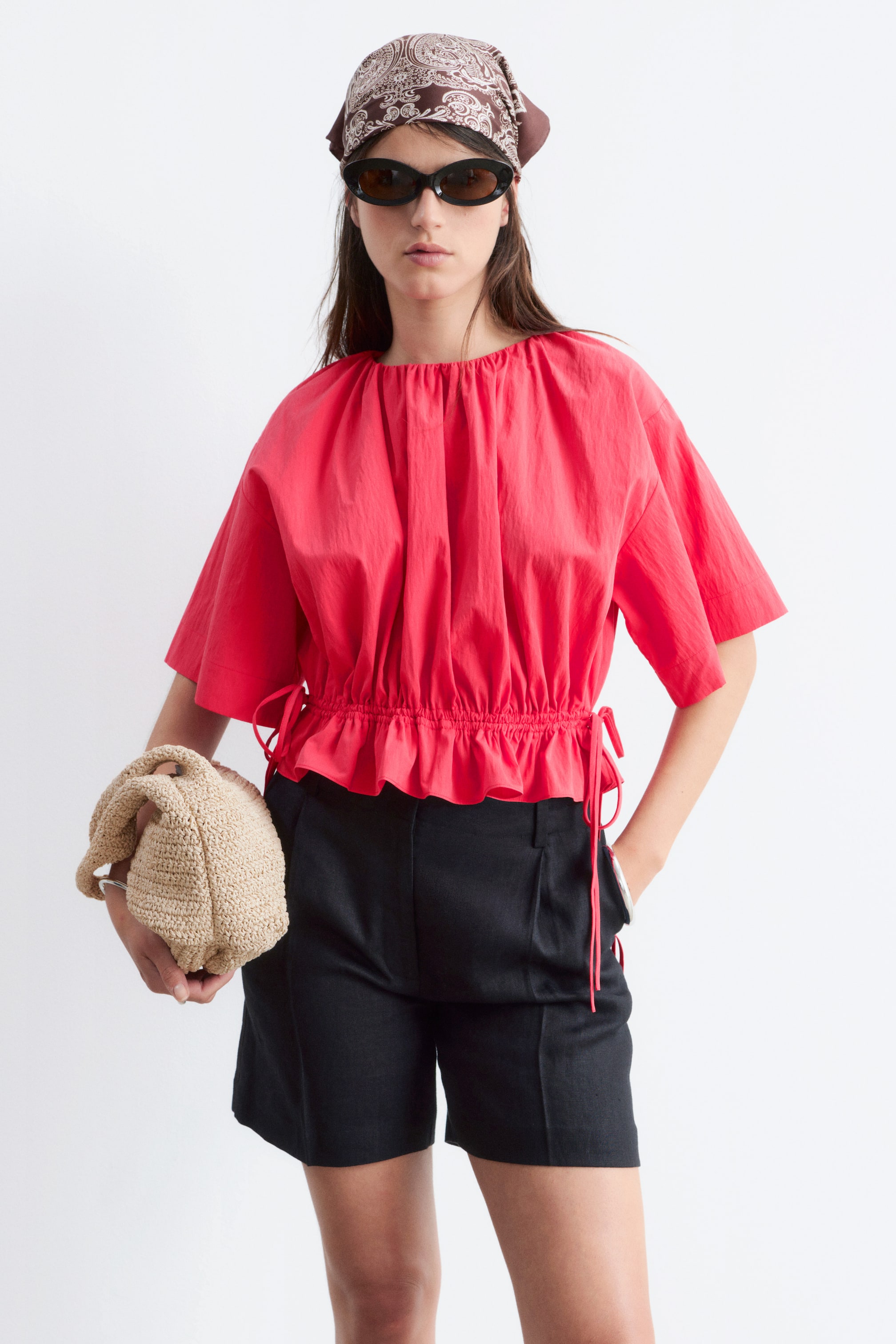 Agrandir l'image: Le haut rouge présente un col rond froncé, des manches courtes et larges, ainsi qu'une taille élastiquée créant un ourlet volanté avec des cordons de serrage visibles sur les côtés.