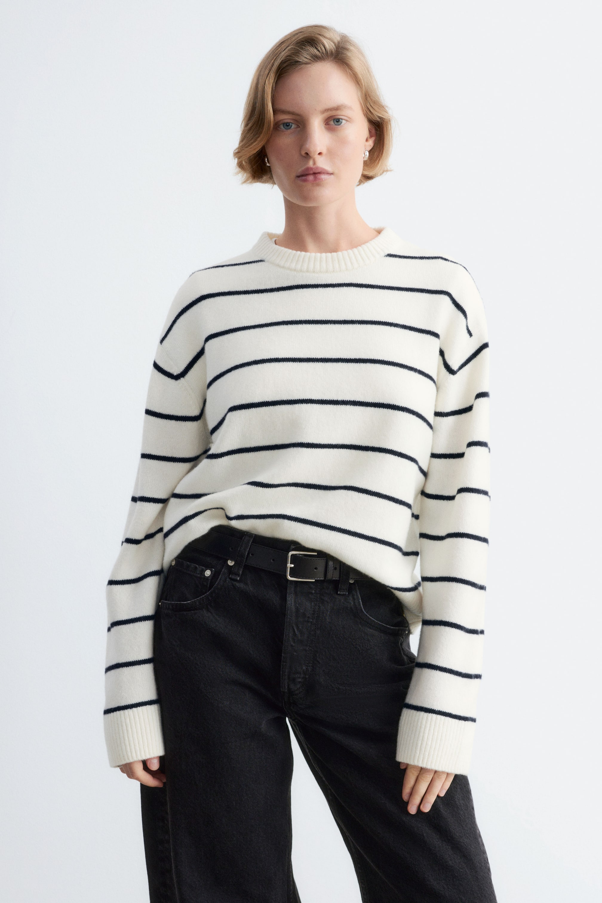 Ver imagem maior: Uma camisola de malha de corte quadrado em creme com riscas horizontais pretas está a ser usada pela modelo. A camisola de manga comprida apresenta gola redonda canelada e punhos canelados, e é combinada com a bainha da frente metida dentro de uns jeans pretos.