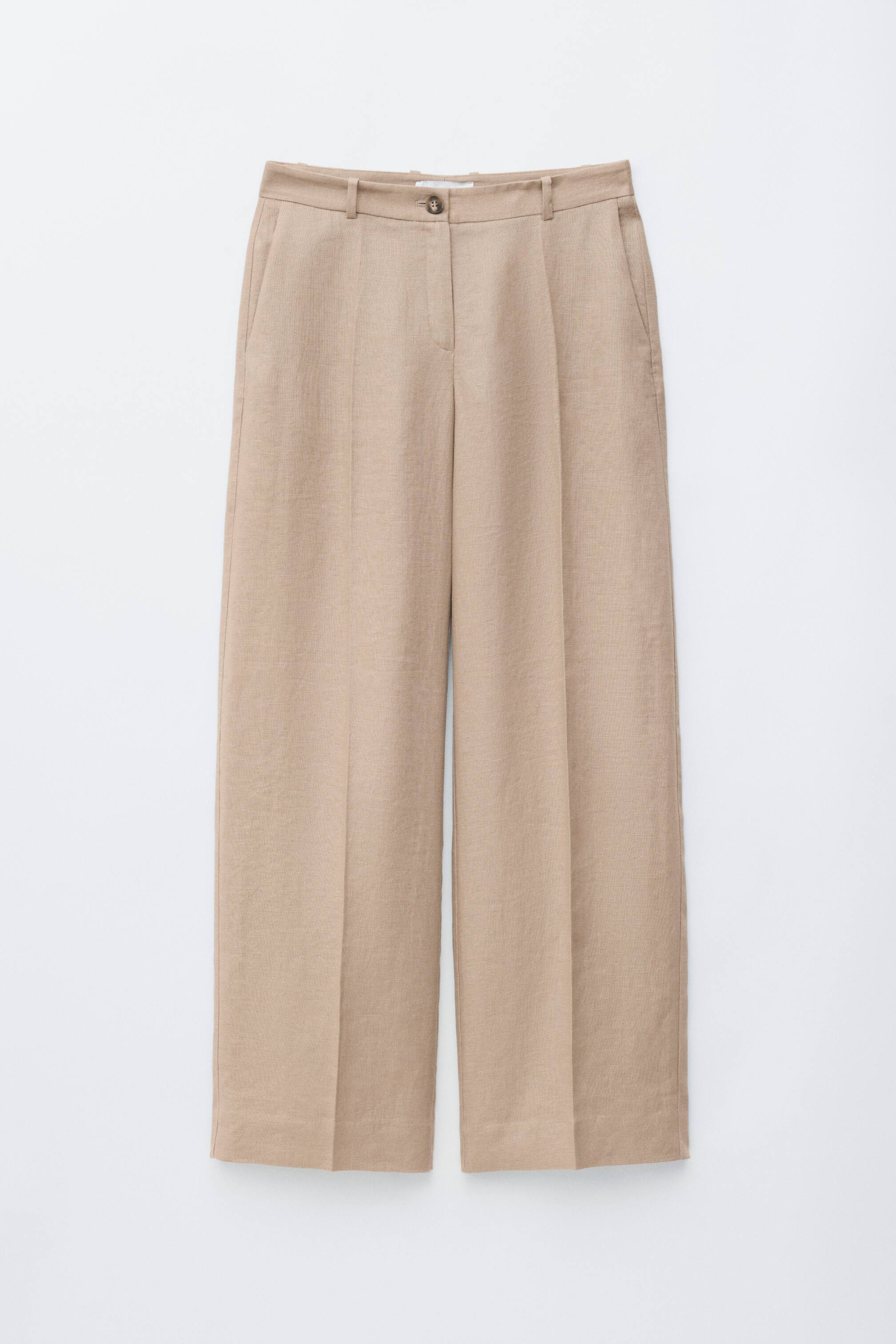 הצגת תמונה גדולה יותר: מכנסי פשתן בצבע בז' בהיר מוצגים מהחזית, עם גזרה רפויה ומותן נמוכה הכוללת סגירת כפתור, רוכסן וכיסים צדדיים אלכסוניים. קפלים לחוצים יורדים לאורך החזית של כל רגל.