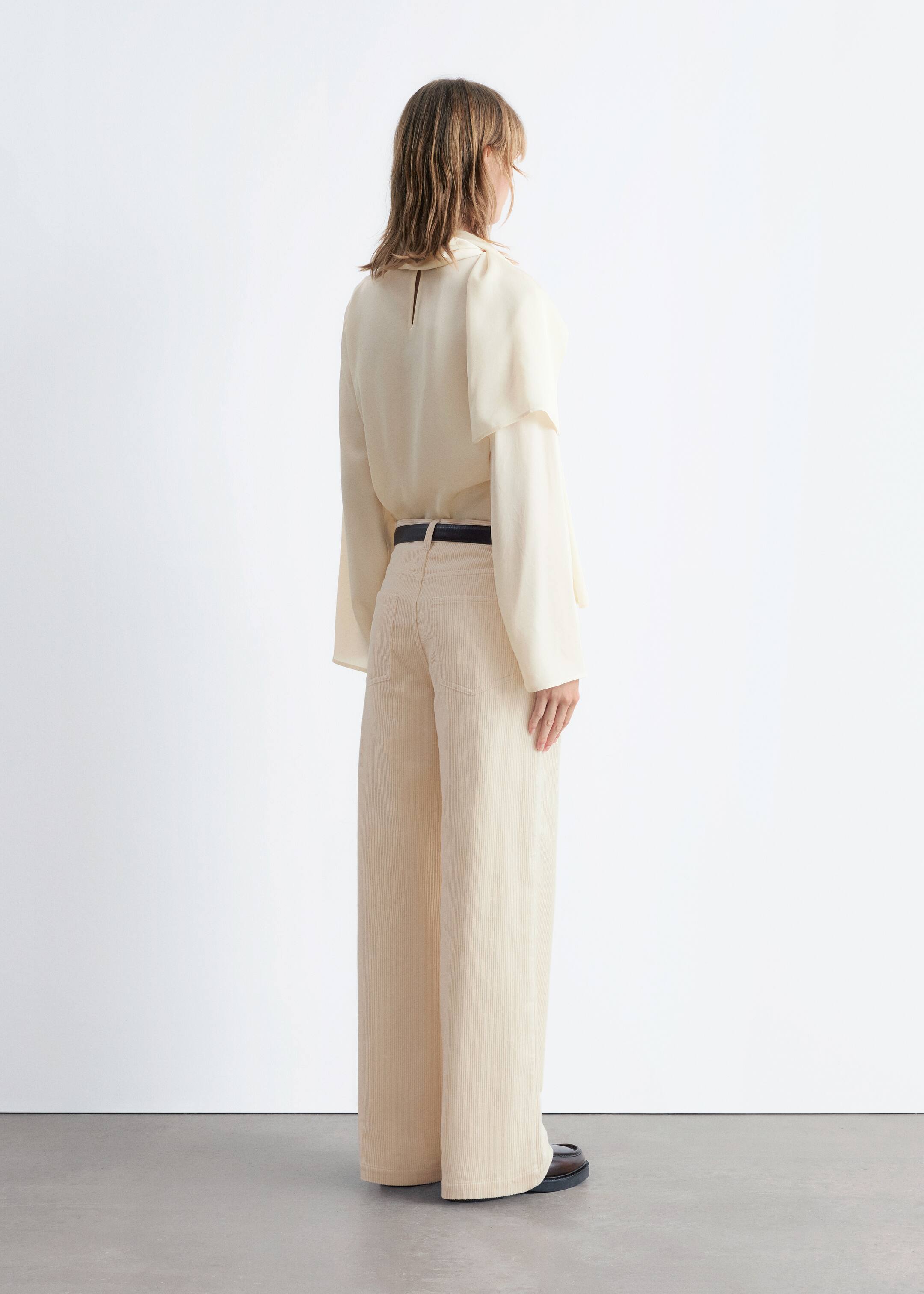 Visualizza immagine più grande: Una blusa color crema, dal taglio ampio, è indossata vista da dietro, caratterizzata da maniche larghe e un dettaglio a strati sulle spalle. È abbinata a pantaloni a gamba larga in velluto a coste beige chiaro, fissati da una cintura sottile scura, con scarpe piatte scure visibili all'orlo.