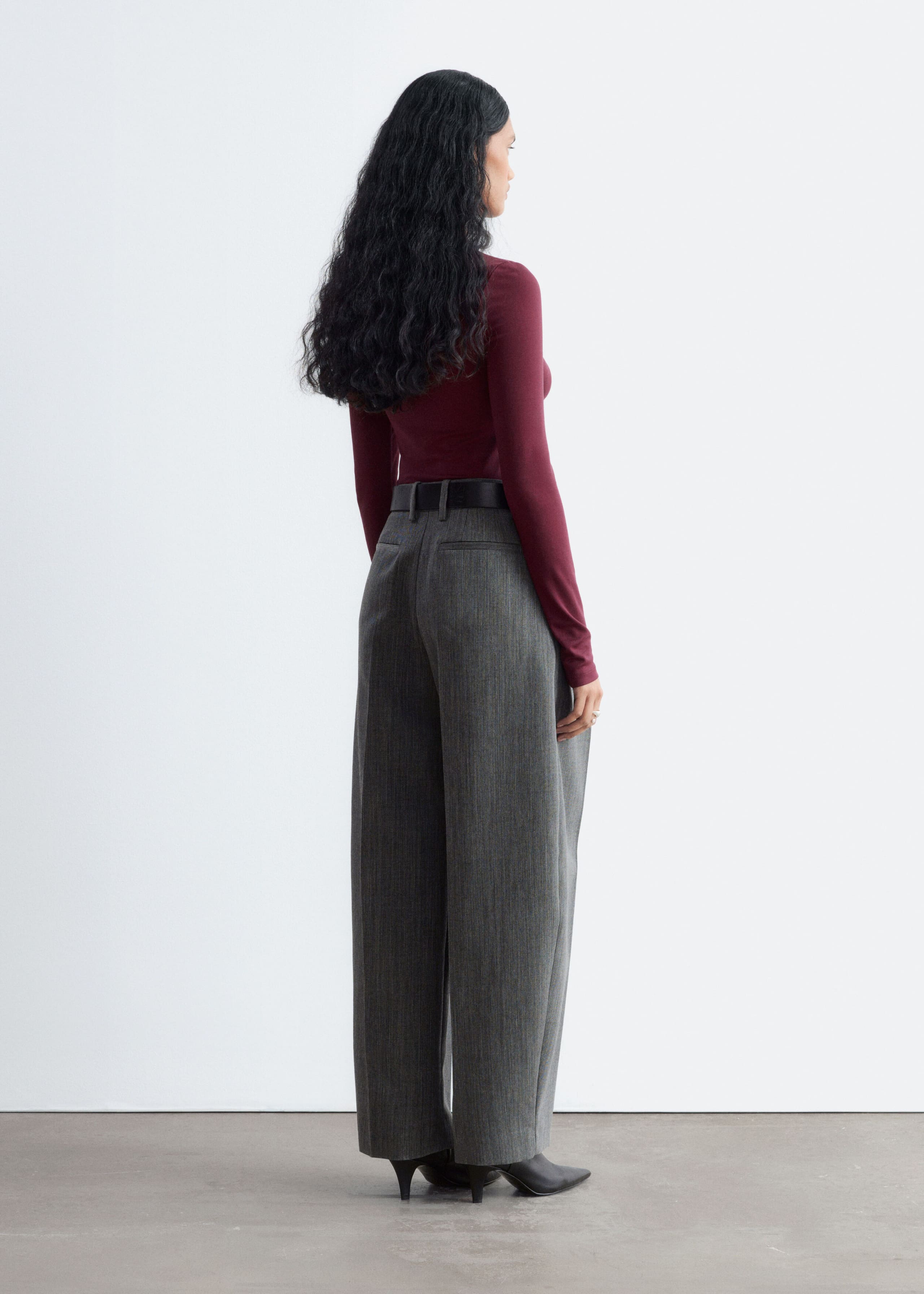 The model is wearing a long-sleeved burgundy jersey top, shown from a three-quarter rear view. The close-fitting top is tucked into high-waisted grey pinstriped trousers styled with a black belt.