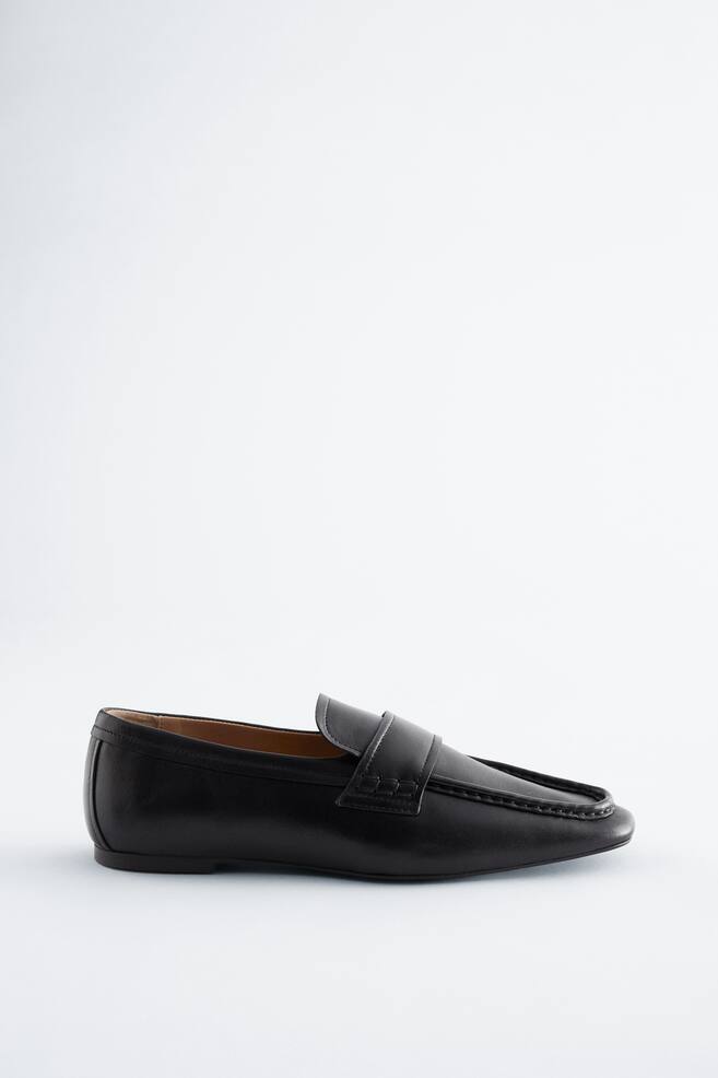 Ein schwarzer Loafer präsentiert ein glattes Finish und ein Penny-Slot-Detail über dem Rist. Er verfügt über eine Mokassin-Zehenkonstruktion mit sichtbaren Nähten und einer niedrigen, flachen Sohle. - 1