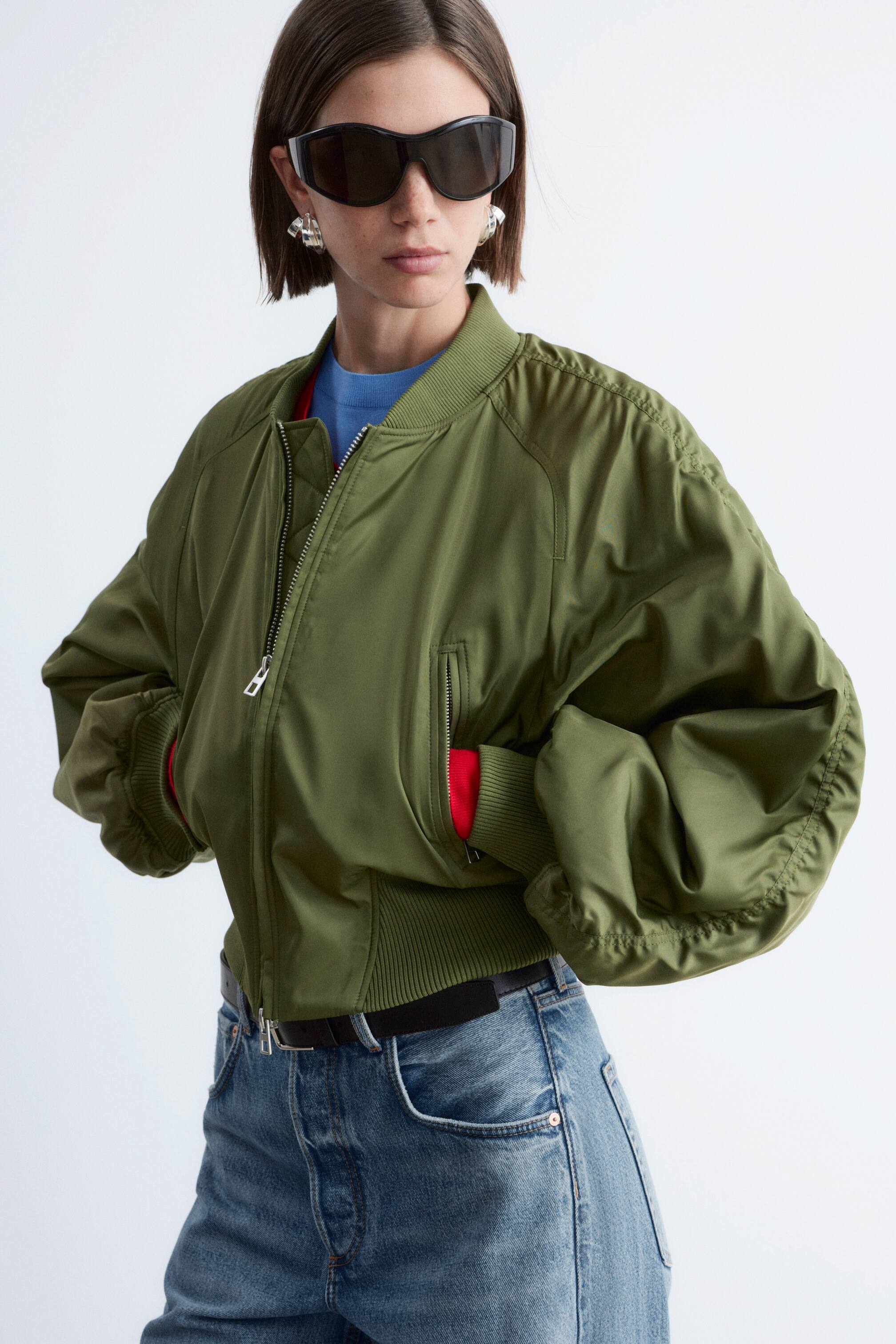 Näytä suurempi kuva: Oliivinvihreä bomber-takki on päällä, siinä on ribattu kaulus, etuvetoketju ja tilavat hihat ribatuilla hihansuilla ja helmalla. Punainen vuori näkyy takin sisäpuolella ja vetoketjulliset sivutaskut, sininen yläosa pilkottaa alta.