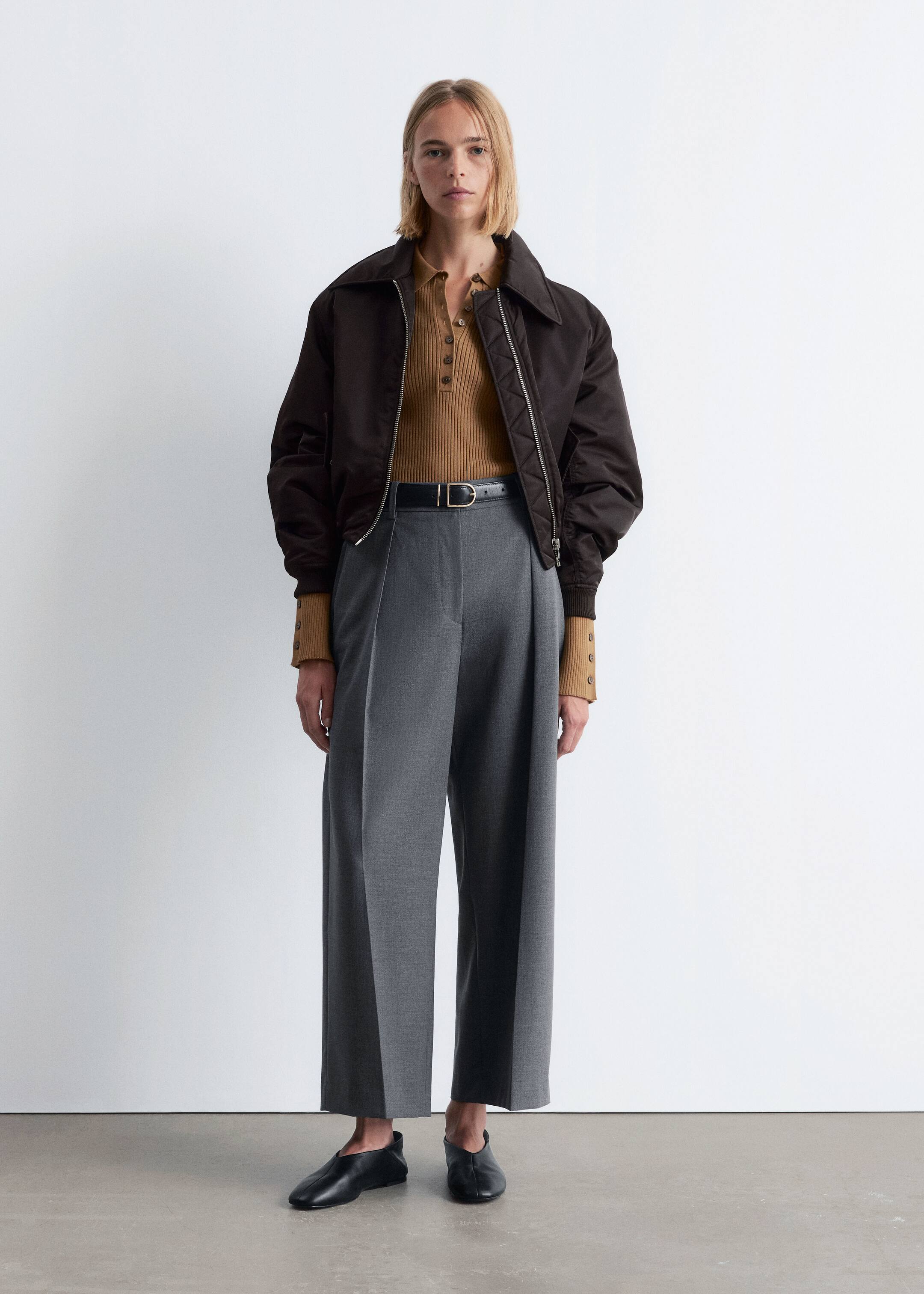 View larger image: An outfit is being presented on a person standing facing forward, featuring a dark brown bomber jacket with a wide collar and quilted zipper placket, layered over a camel ribbed polo top with a three-button placket and buttoned cuffs. The ensemble includes grey wide-leg pleated trousers with a cropped hem, cinched by a black belt with a gold-tone D-ring buckle, and black slip-on shoes.