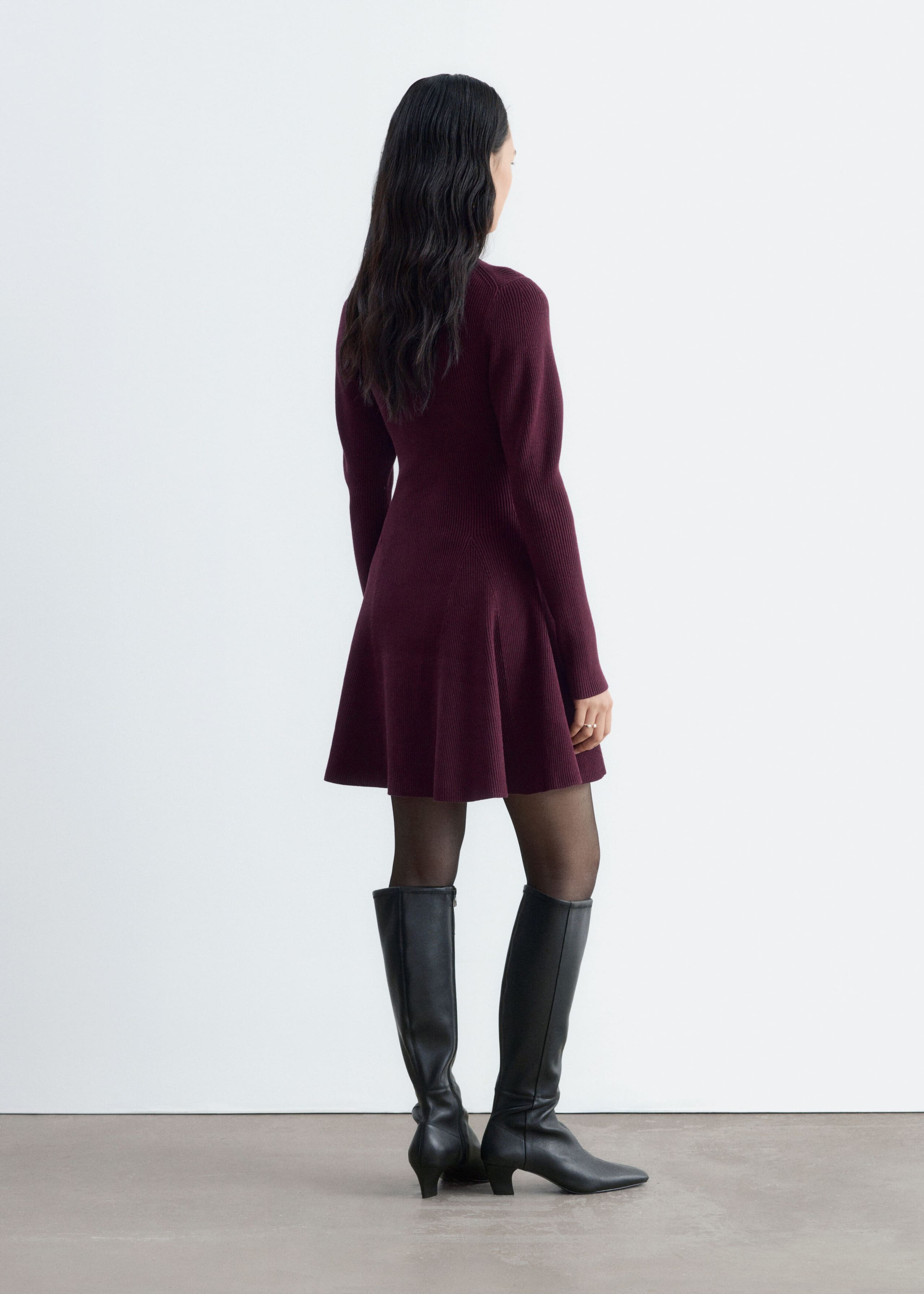 A burgundy ribbed-knit dress is worn by the model, highlighting its long sleeves, fitted bodice, and flared paneled skirt from a three-quarter back angle. The dress is styled with sheer black tights and black knee-high leather boots.