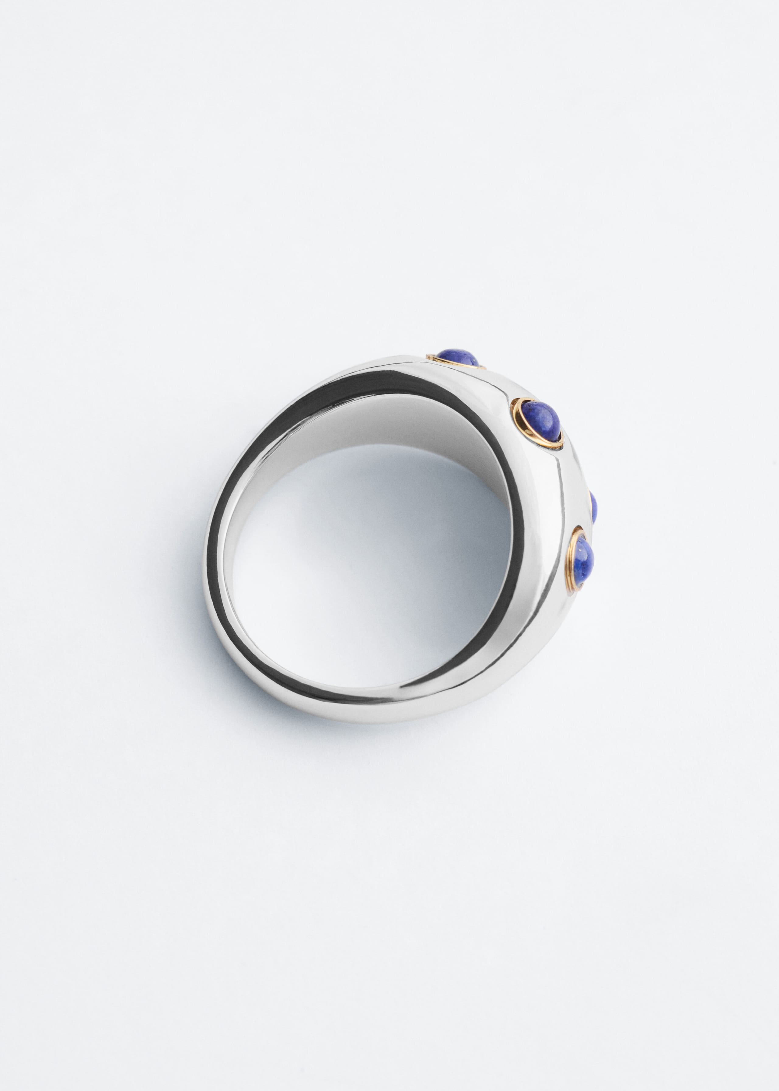 Ein skulpturaler, polierter Ring in Silberoptik liegt schräg und präsentiert sein dickes, geschwungenes Band sowie die kreisförmige Öffnung. Die Außenseite ist mit kleinen, runden blauen Cabochon-Steinen versehen, die in goldfarbenen Fassungen gefasst sind.