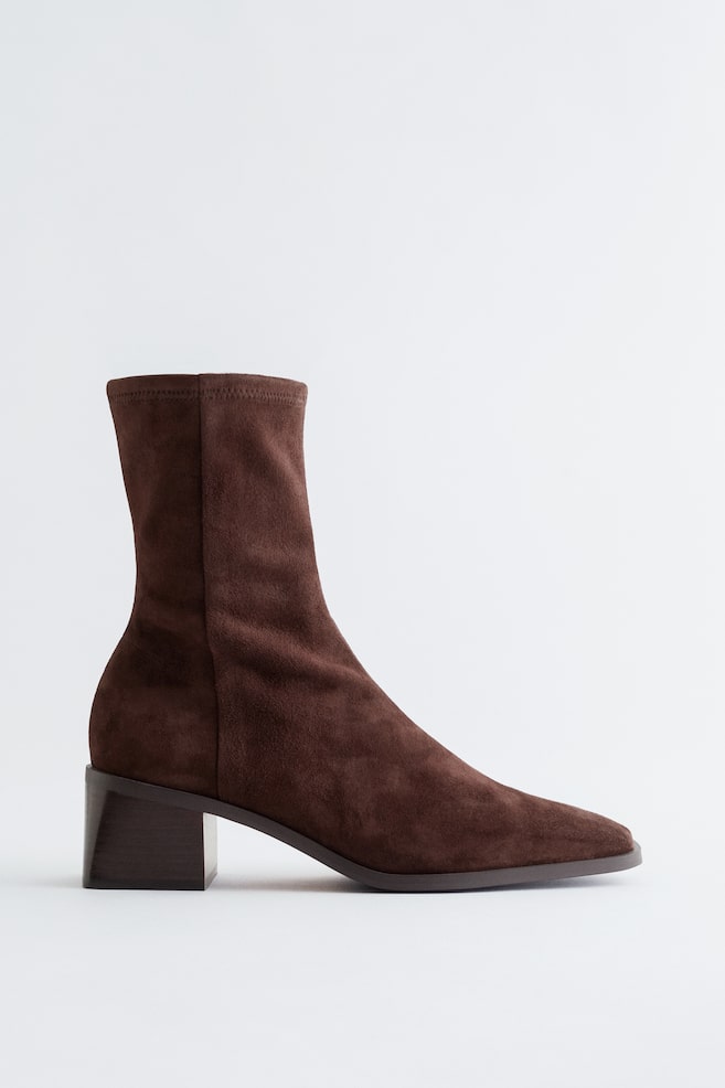 A dark brown suede-textured sock boot is being displayed, featuring a squared toe, a chunky block heel, and a prominent topstitched center seam running up the front of its shaft. - 1