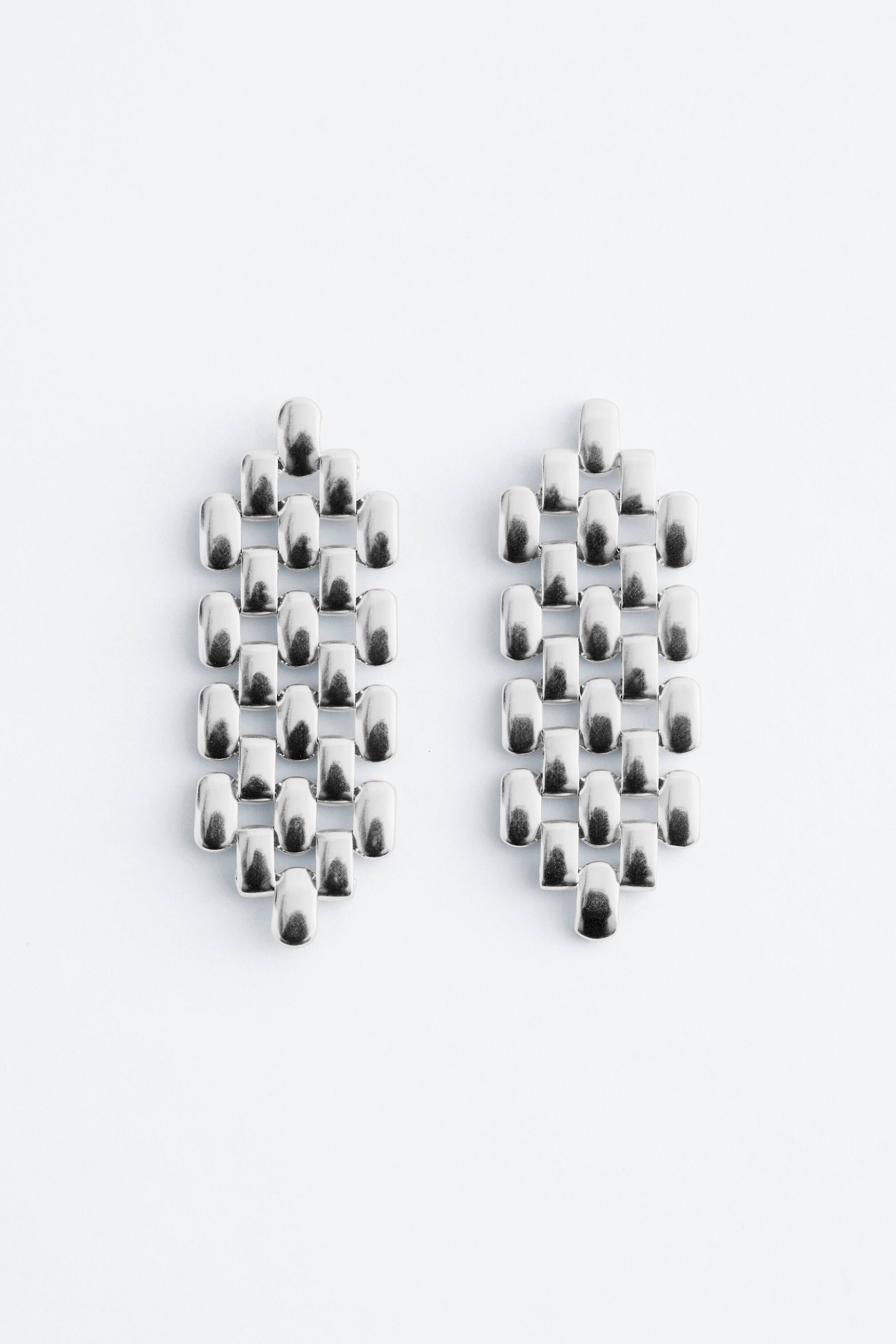 View larger image: A pair of silver-toned pendant earrings featuring an architectural chain pattern, crafted from numerous interlinked, rounded rectangular segments, presenting an elongated, tapering shape with a smooth, reflective finish.