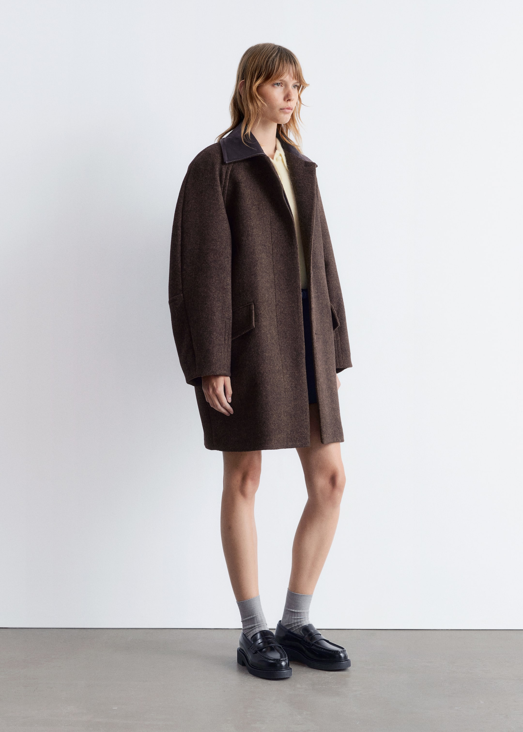 View larger image: A dark brown, textured coat is being presented from a right-facing angle. The oversized coat features a contrasting dark brown spread collar and two visible flap pockets on the front, extending to mid-thigh length, and is being worn open over a light yellow top.