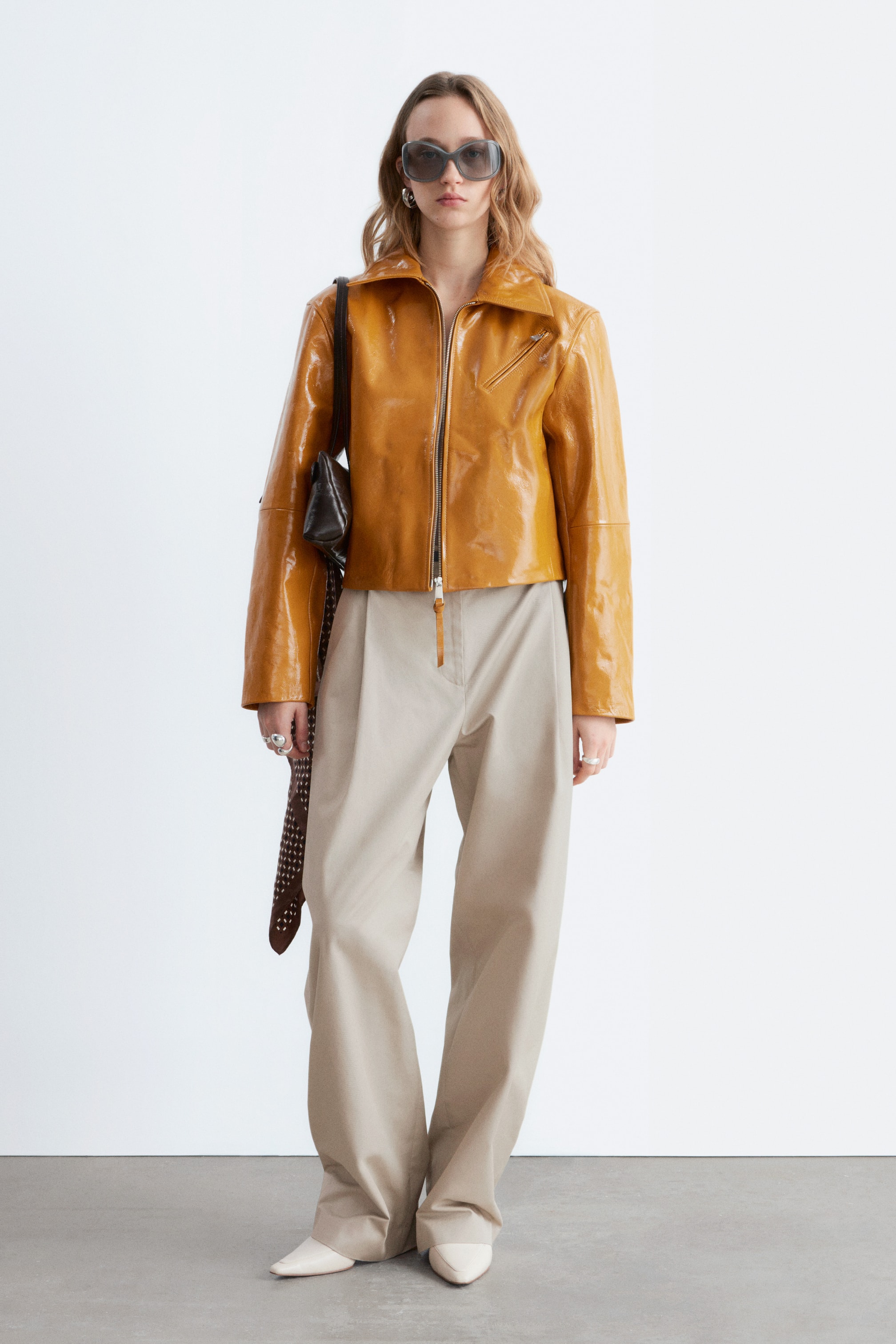 Visa större bild: Ljusbeige högmidjade byxor bärs av modellen. De har raka, avslappnade ben med skarpa pressveck framtill och en synlig frontstängning.