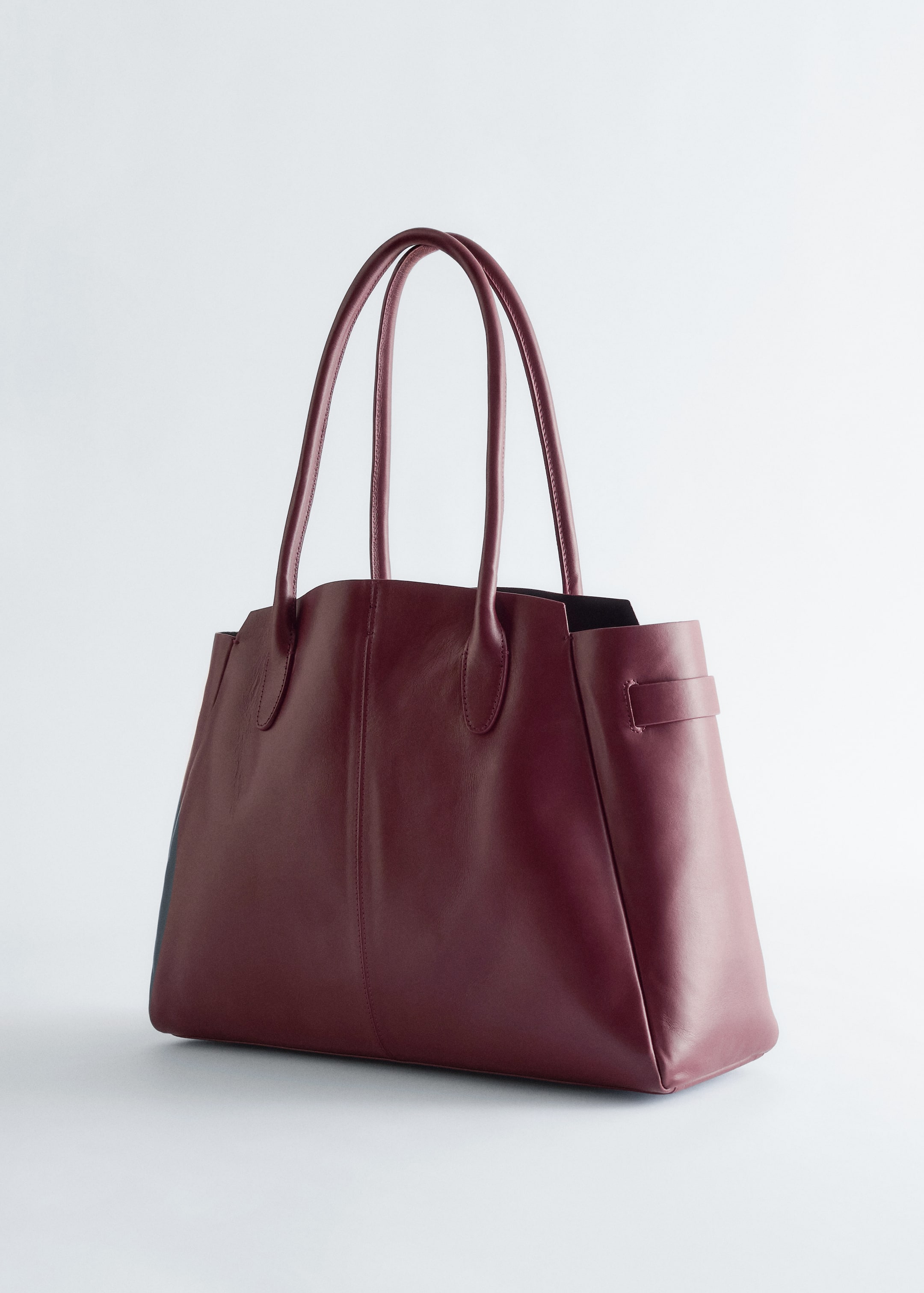 View larger image: A deep red tote bag is positioned at a front-right angle, featuring a smooth, structured exterior with two top handles, contrasting dark-toned side gussets, and a small band detail visible on the right side.