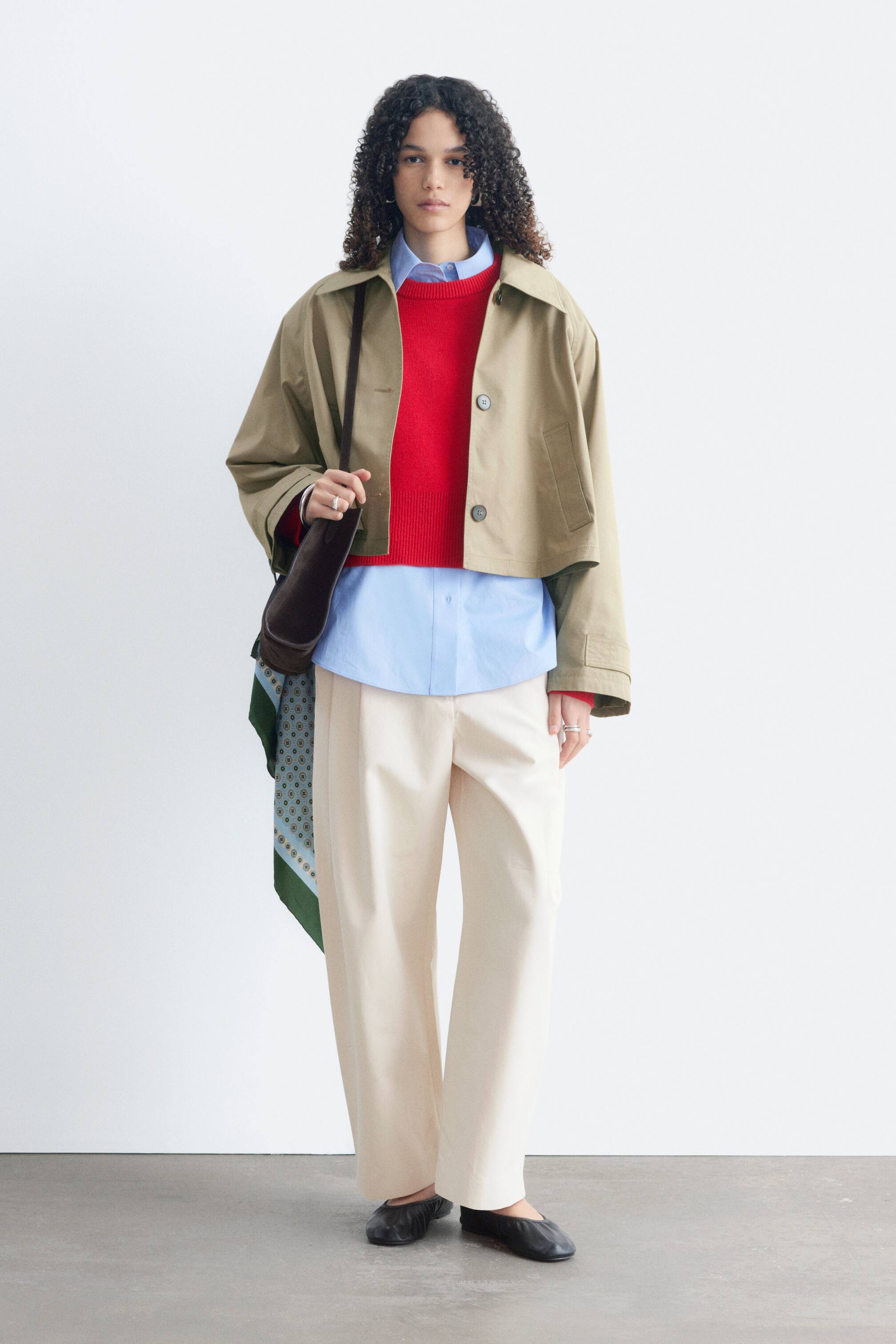 Grotere afbeelding bekijken: Een khaki cropped jasje met een losse pasvorm, knoopsluiting, paspelzakken en mouwtabbladen met knopen wordt gedragen door het model. Het is gelaagd over een rode trui en een lichtblauw hemd, met een donkerbruine schoudertas die over het jasje gedragen wordt.