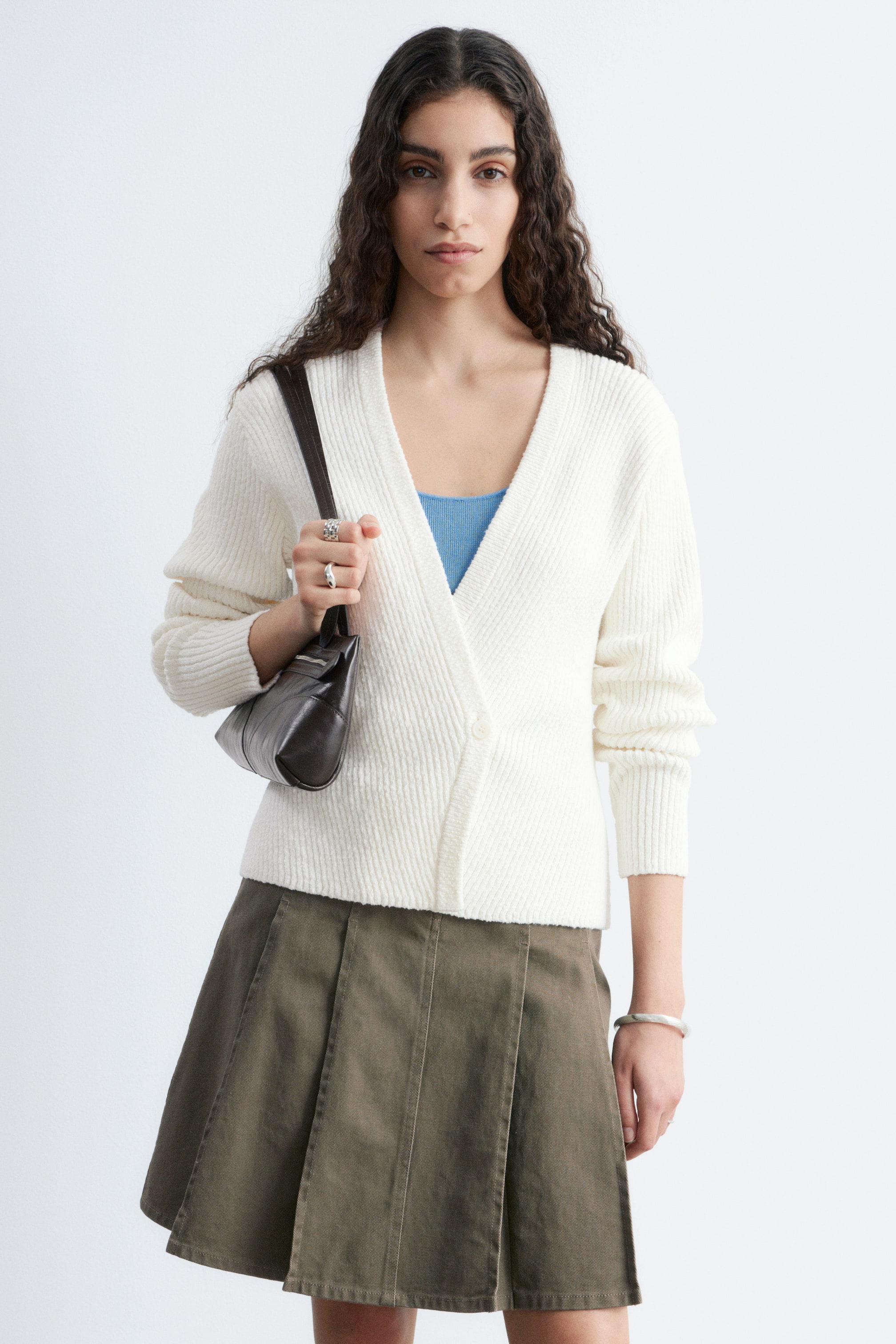 הצגת תמונה גדולה יותר: קרדיגן מעטפת בצבע קרם נלבש, מציג מרקם סריג עם צלעות, מחשוף V ושרוולים ארוכים עם חפתים מצולעים. הוא נסגר עם כפתור אחד בצד ימין, מעל חולצה כחולה.