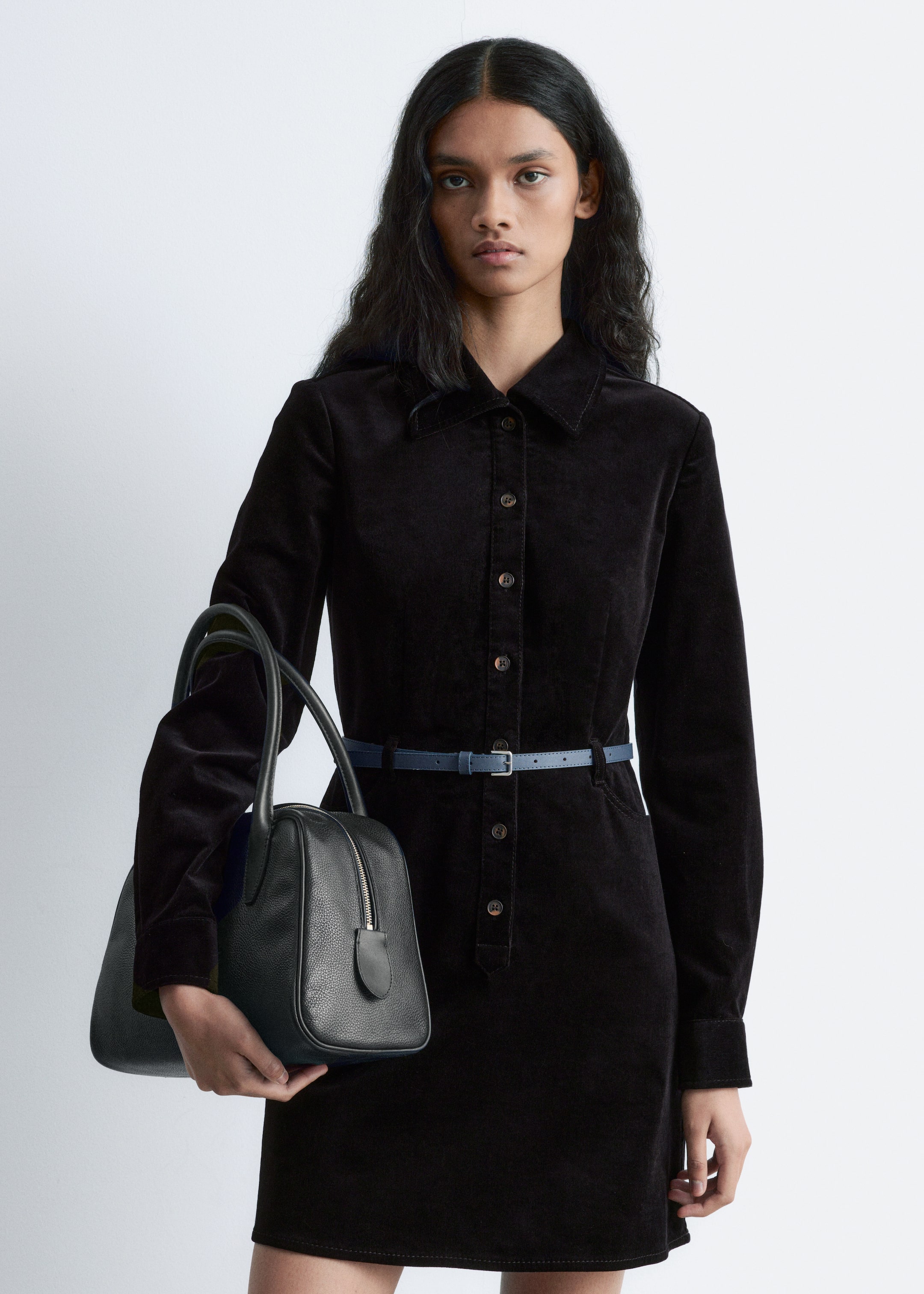 Ver imagem maior: Um vestido-camisa de veludo cotelê preto, com gola, mangas compridas e fecho completo de botões na frente, está a ser apertado na cintura por um cinto fino de couro azul com uma fivela prateada. Uma mala estruturada de couro preto com textura de seixos, com alça superior e fecho de correr na parte superior, está a ser segurada ao lado.