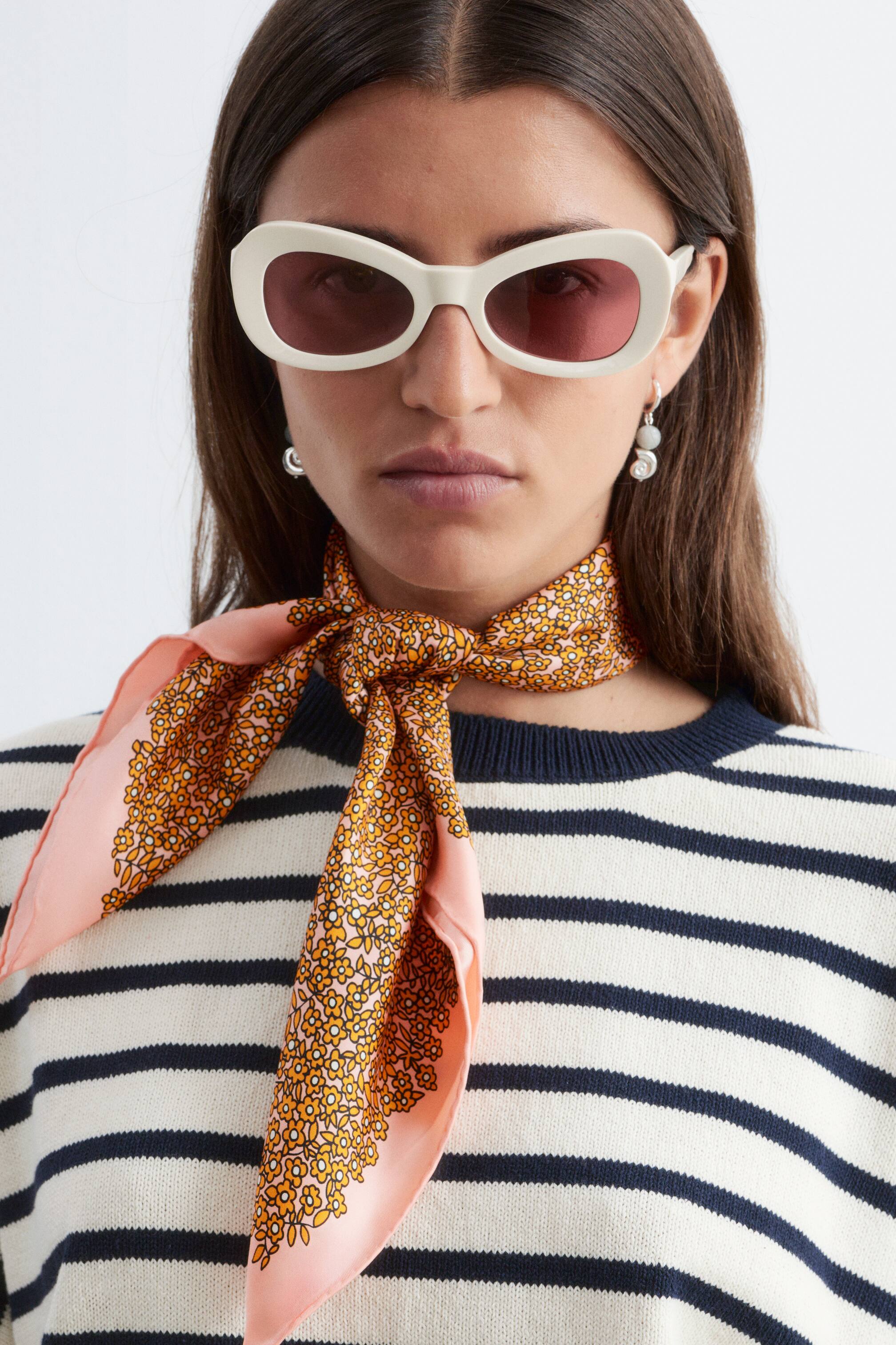 Zobraziť väčší obrázok: Štvorcová šatka je uviazaná okolo krku, pričom zobrazuje svetloružový okraj a hustý vzor malých oranžových a žltých kvetov. Je zaviazaná vpredu a jej konce sú prehodené cez biely a tmavomodrý pruhovaný sveter.