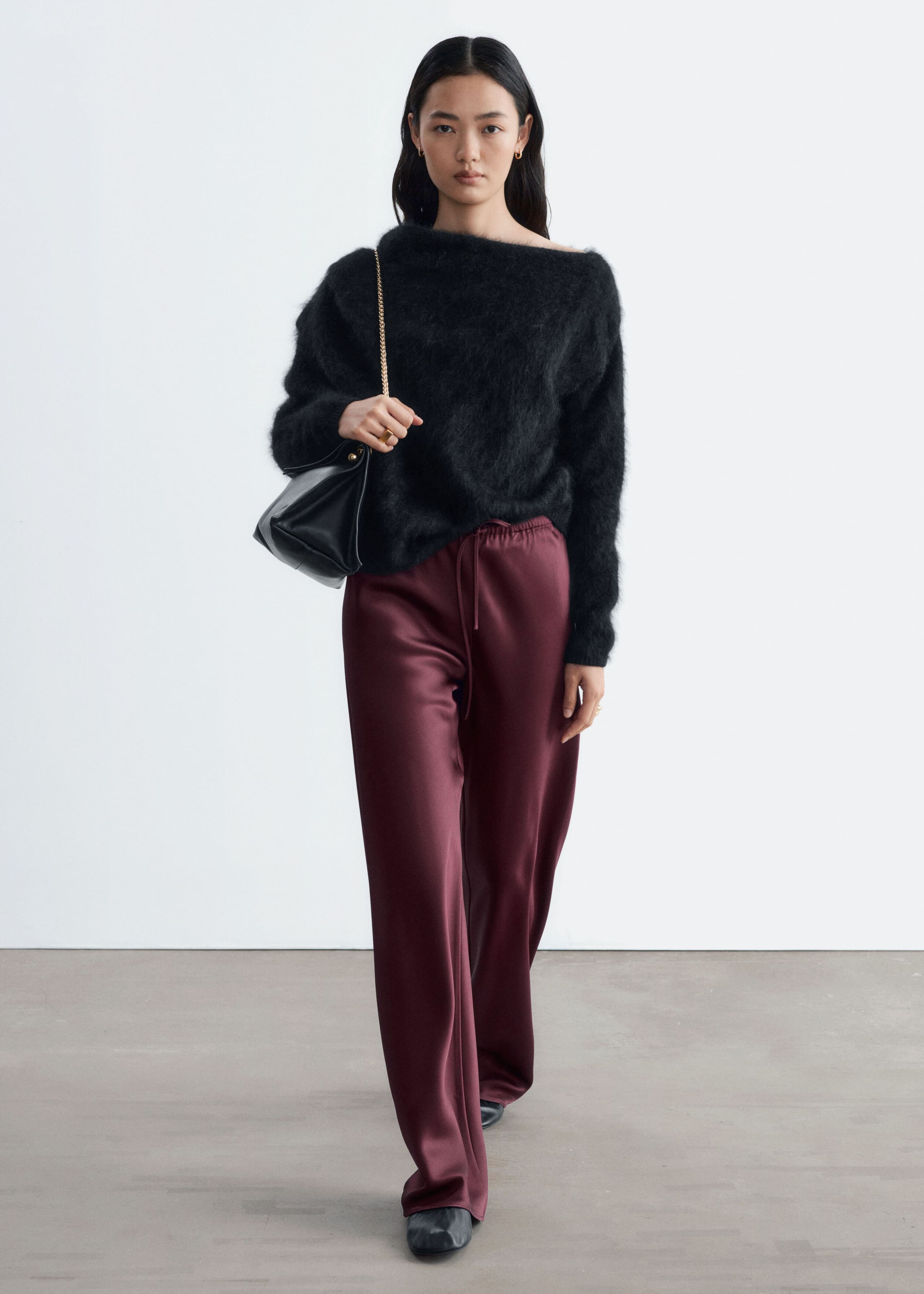 The model is wearing burgundy satin trousers with a relaxed fit, an elastic drawstring waistband, and wide legs. The trousers are styled with a black fuzzy off-the-shoulder sweater and a black shoulder bag.