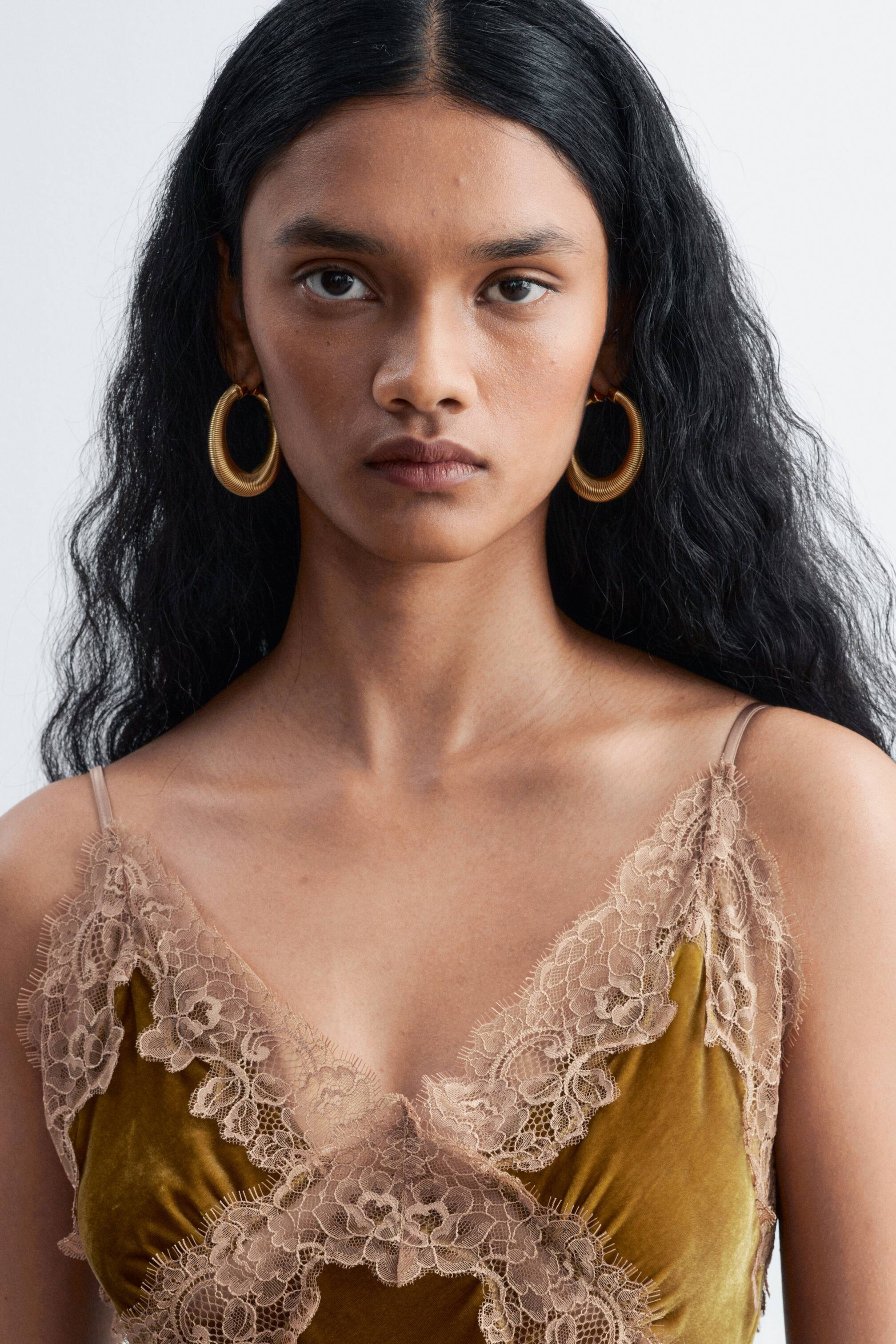 View larger image: An olive-green velvet slip dress featuring delicate beige lace trim along the V-neckline is being worn by the model, paired with chunky, textured gold hoop earrings.