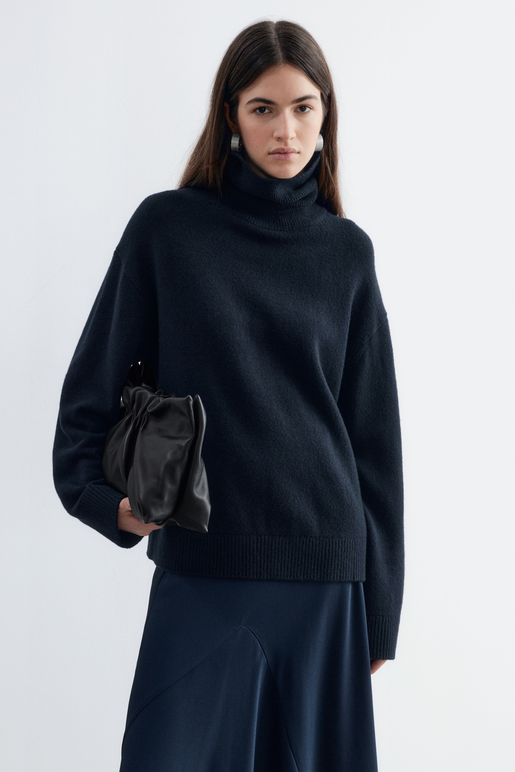 Größeres Bild anzeigen: Ein dunkelmarineblauer Rollkragenpullover aus Woll-Baumwoll-Mischung mit lockerem Schnitt, hohem Rollkragen und Rippbündchen wird von dem Model getragen. Der Pullover wird mit einem dunkelblauen Midirock kombiniert, und unter dem Arm des Models wird eine geraffte schwarze Leder-Clutch getragen.