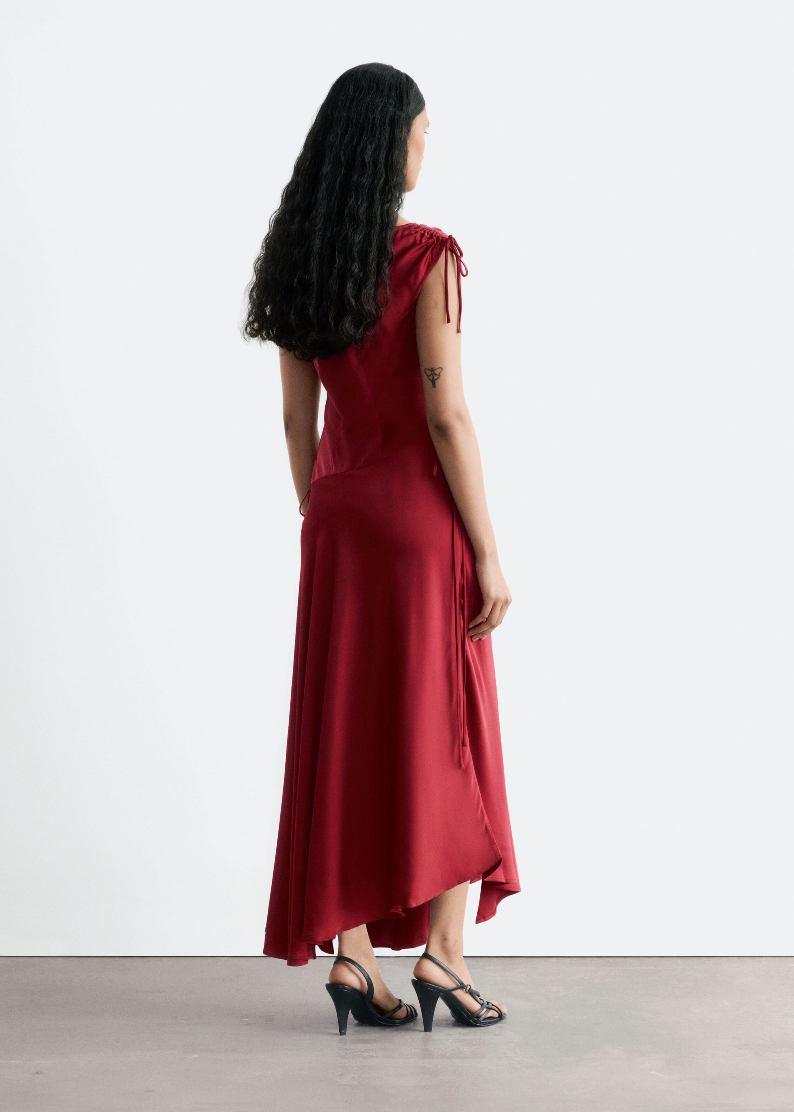 A dark red satin draped dress is being worn by the model, shown from a three-quarter rear view. The dress features drawstring detailing at the shoulder, a gathered dropped waist, and an asymmetrical flowing hemline.