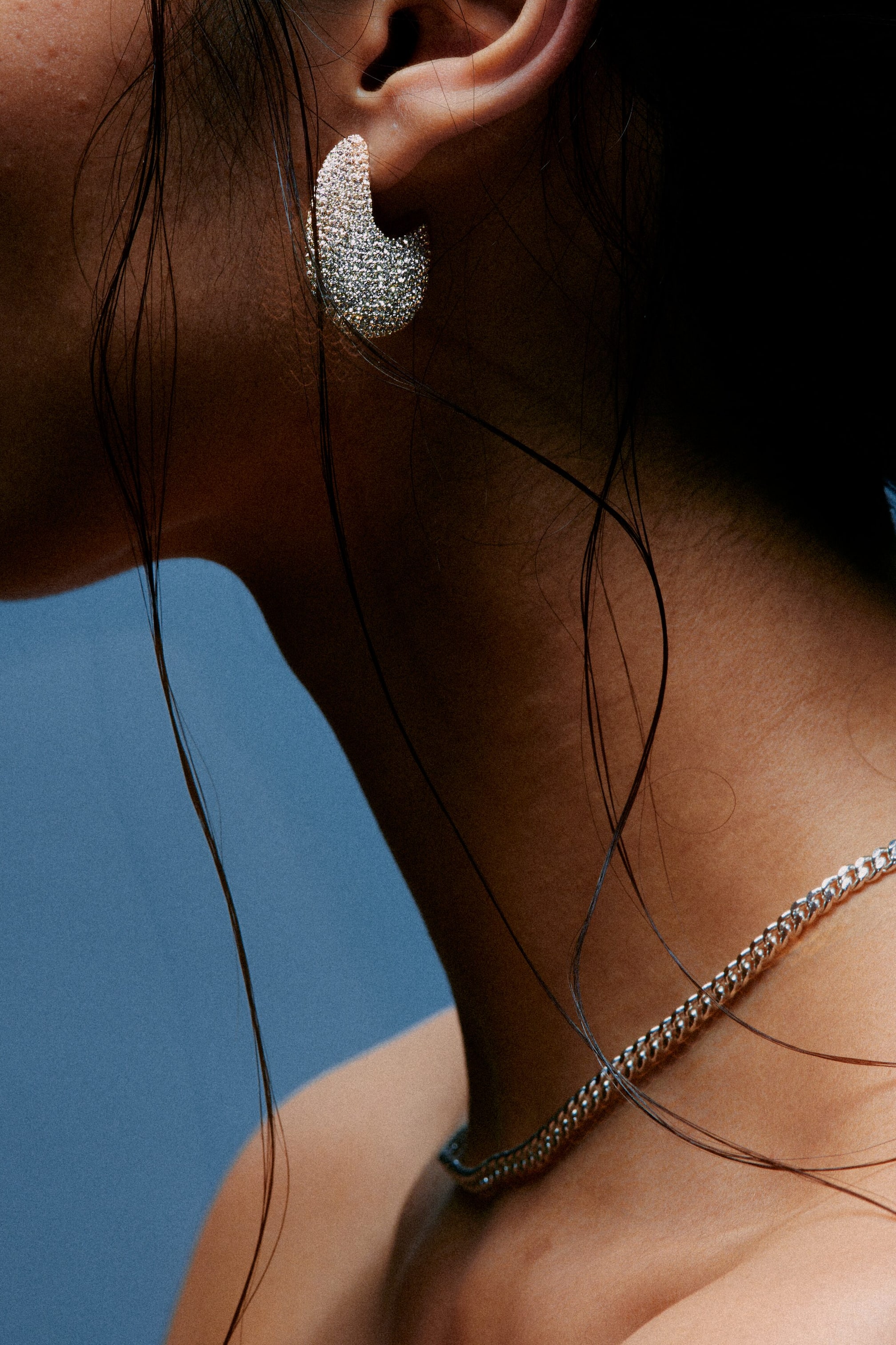 View larger image: A C-shaped earring, encrusted with sparkling pavé stones, is being worn on an earlobe. A silver-toned curb chain necklace is resting around the neck.