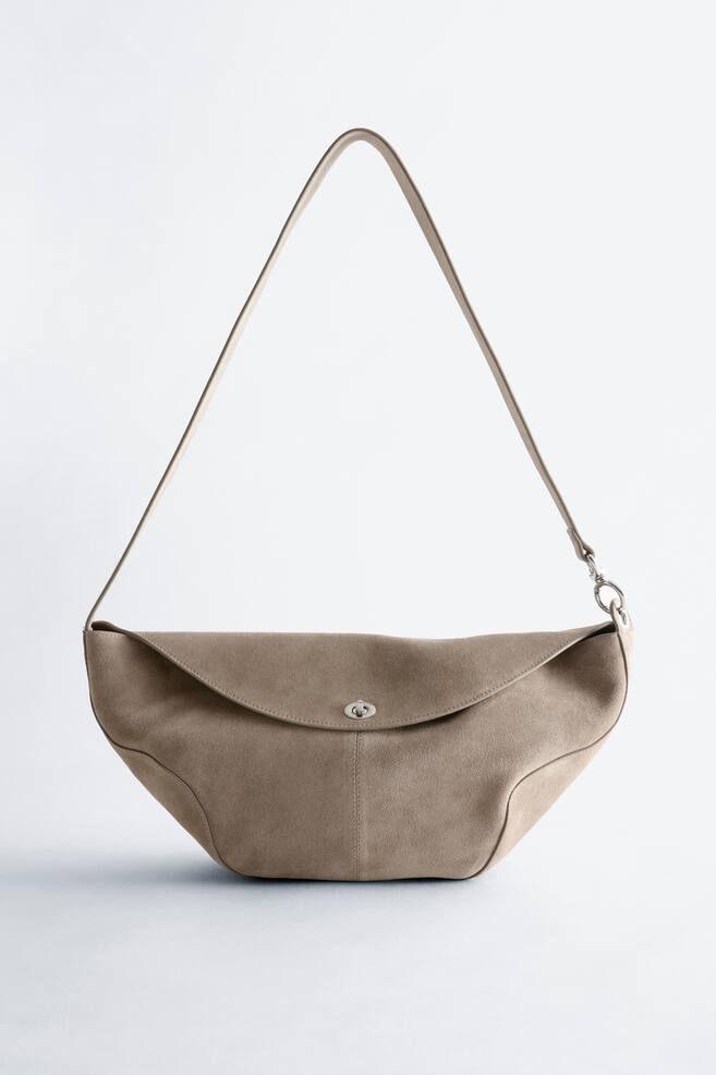 Eine beige Wildleder-Umhängetasche präsentiert sich aus der Vorderansicht mit einer skulpturalen, geschwungenen Silhouette. Sie verfügt über eine abgerundete Klappe, die durch ein silberfarbenes Metall-Drehschloss gesichert wird, mit einem einzelnen dünnen Riemen, der an den Seiten befestigt ist. - 1
