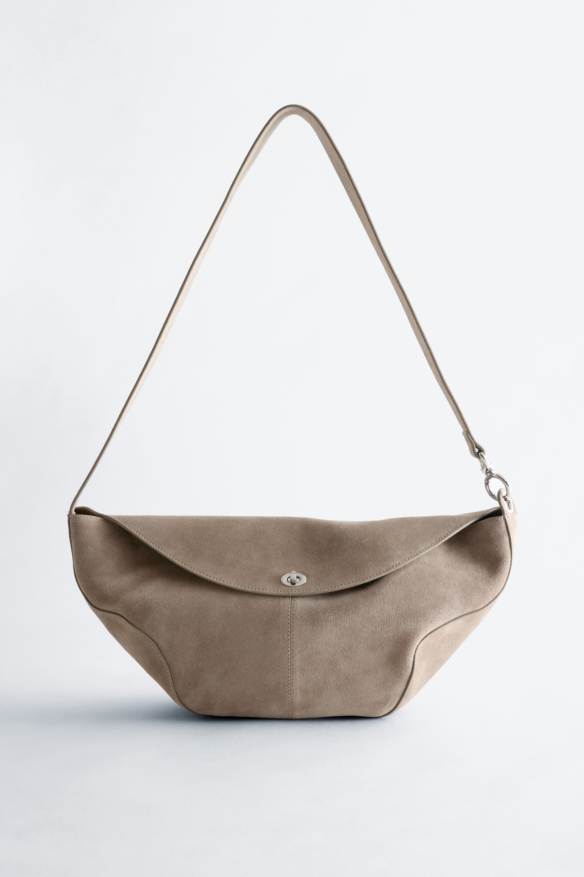 View larger image: A beige suede crossbody bag showcases a sculptural, curved silhouette from a front view. It features a rounded flap secured by a silver-toned metal twist lock, with a single thin strap attaching to the sides.