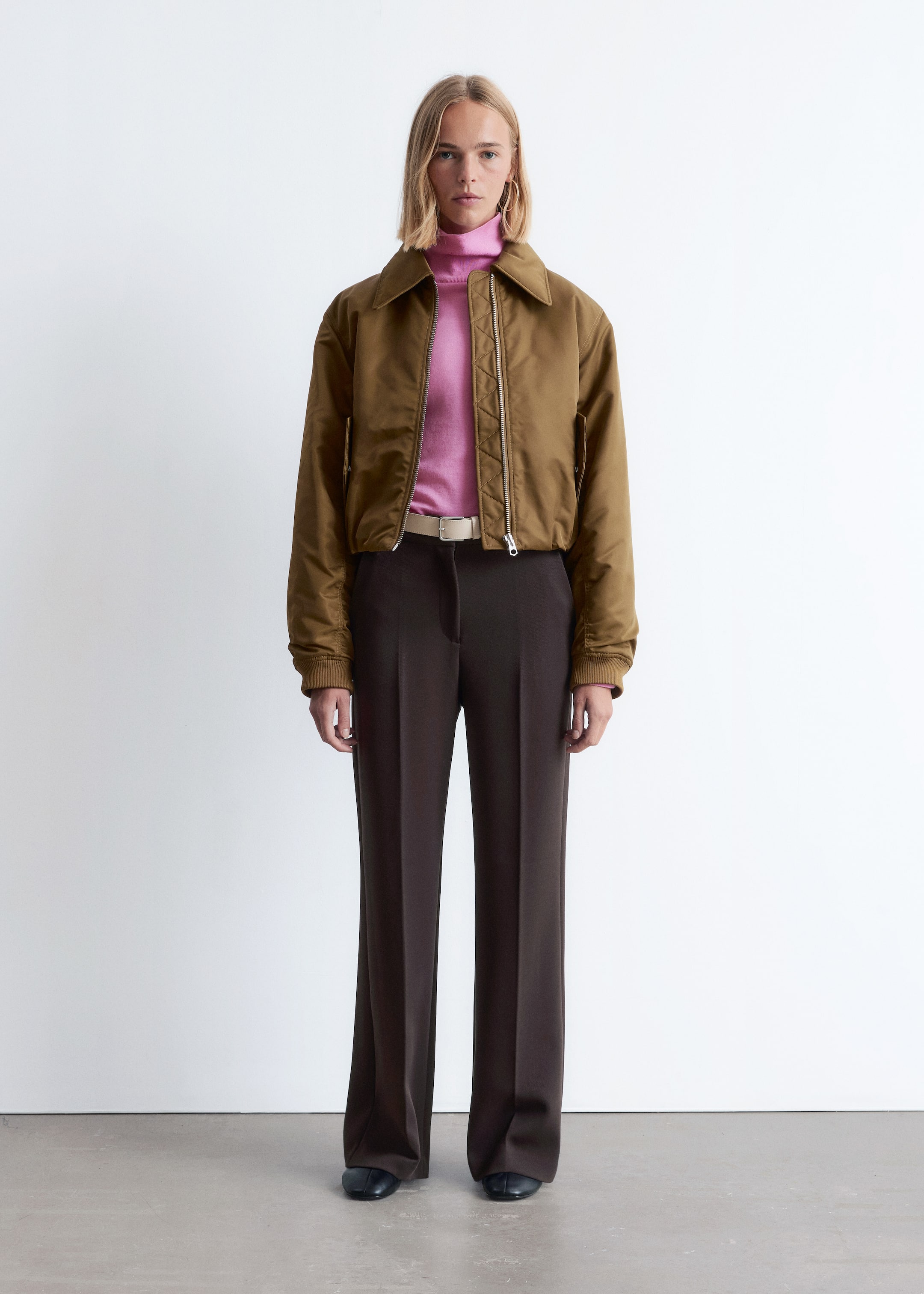 Agrandir l'image: Une tenue complète est présentée de face, mettant en vedette un blouson bomber court olive-brun avec un col pointu, une fermeture éclair argentée à l'avant et des poignets côtelés, superposé à un pull à col roulé rose. Un pantalon large à taille haute de couleur brun foncé avec des plis à l'avant est porté, maintenu par une ceinture beige clair avec une boucle rectangulaire, avec des chaussures plates foncées complétant l'ensemble.