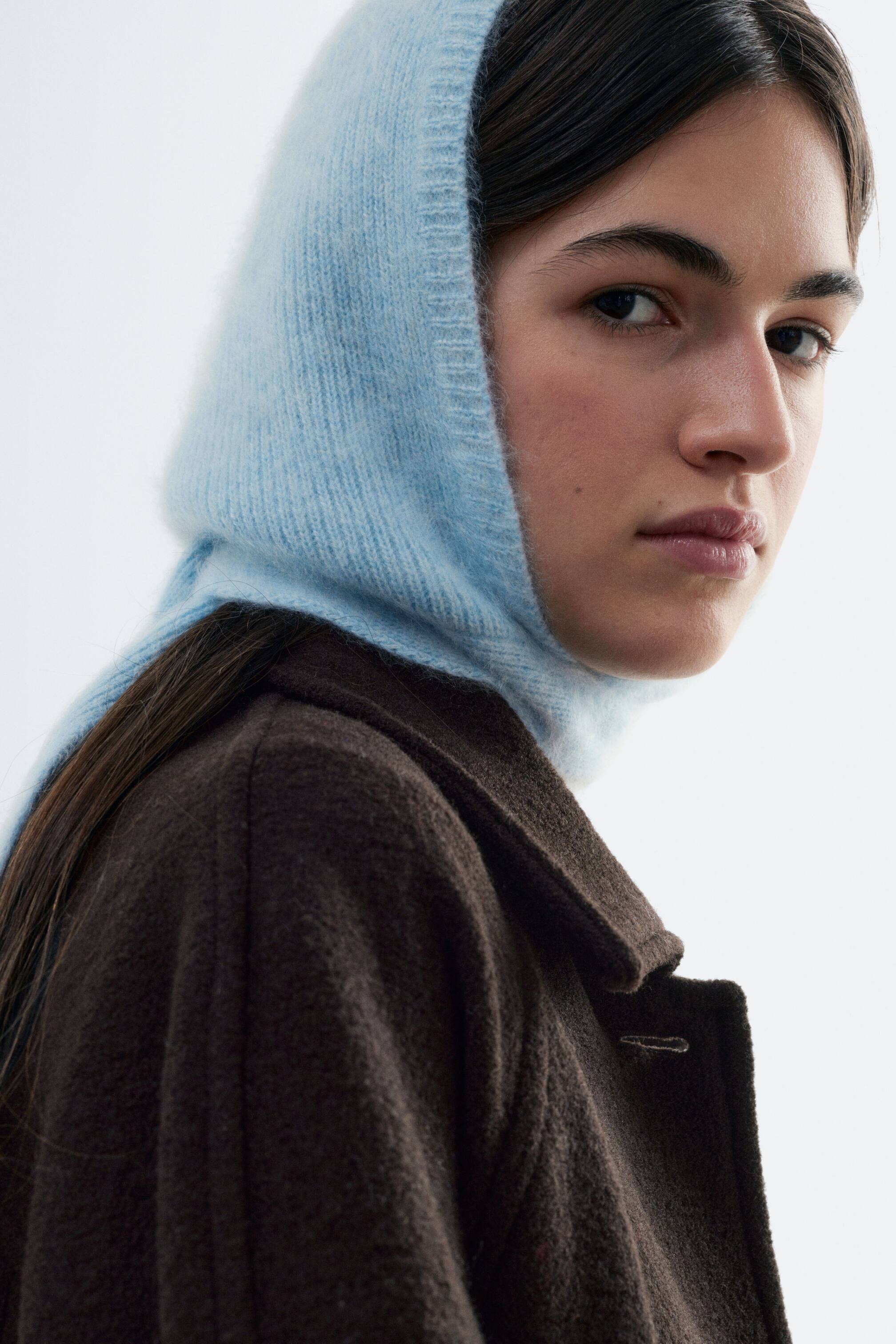 View larger image: The model is wearing a light blue brushed cashmere bonnet, covering the head and ears, with a ribbed trim framing the face. The textured knit bonnet extends down the back of the neck and is layered with a dark brown wool coat.