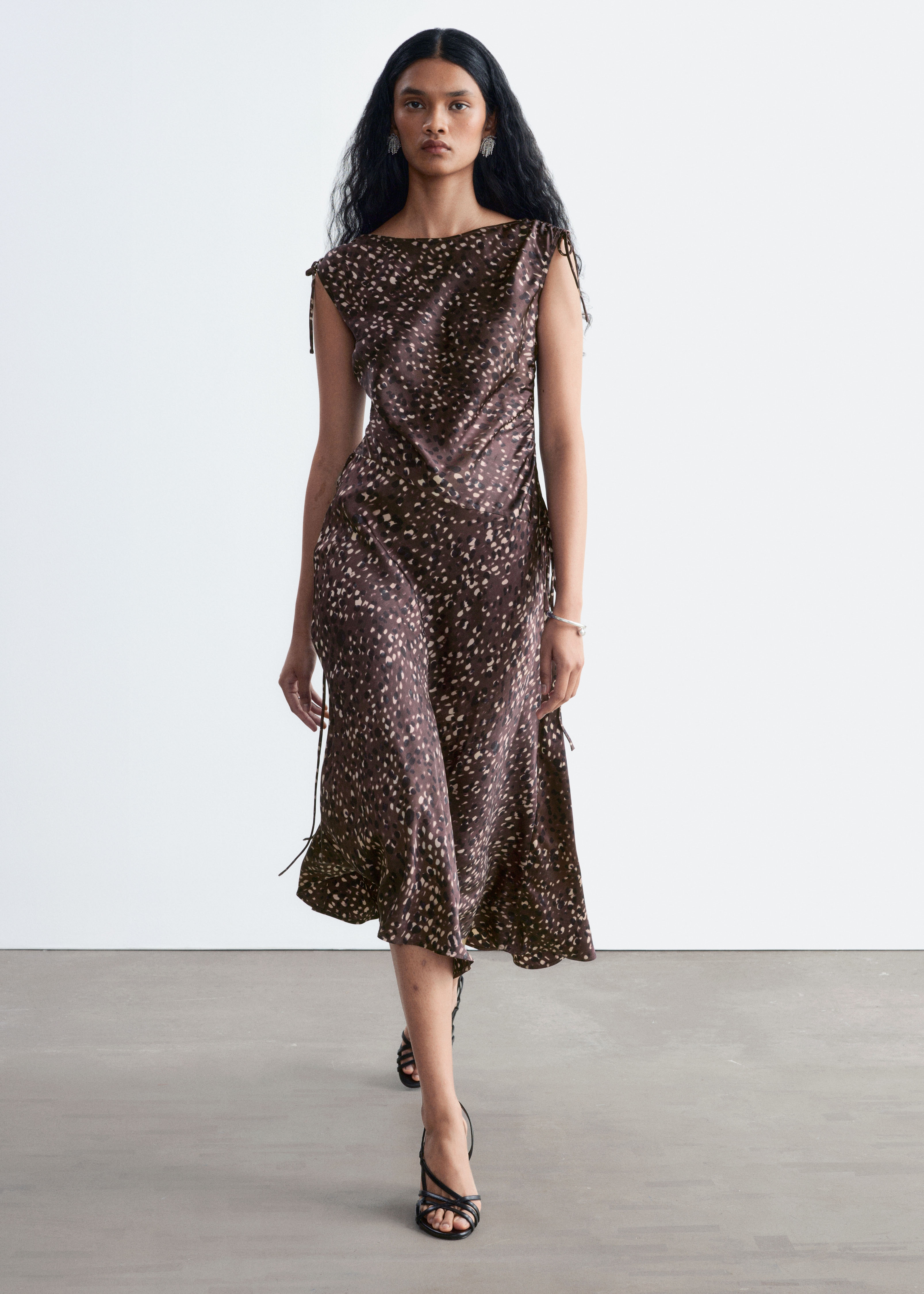 Um vestido midi acetinado sem mangas, com estampado abstrato salpicado castanho, é apresentado de frente, com alças ajustáveis por cordão e corpete drapeado. O vestido exibe uma cintura assimétrica rebaixada que desce para uma bainha assimétrica evasé.