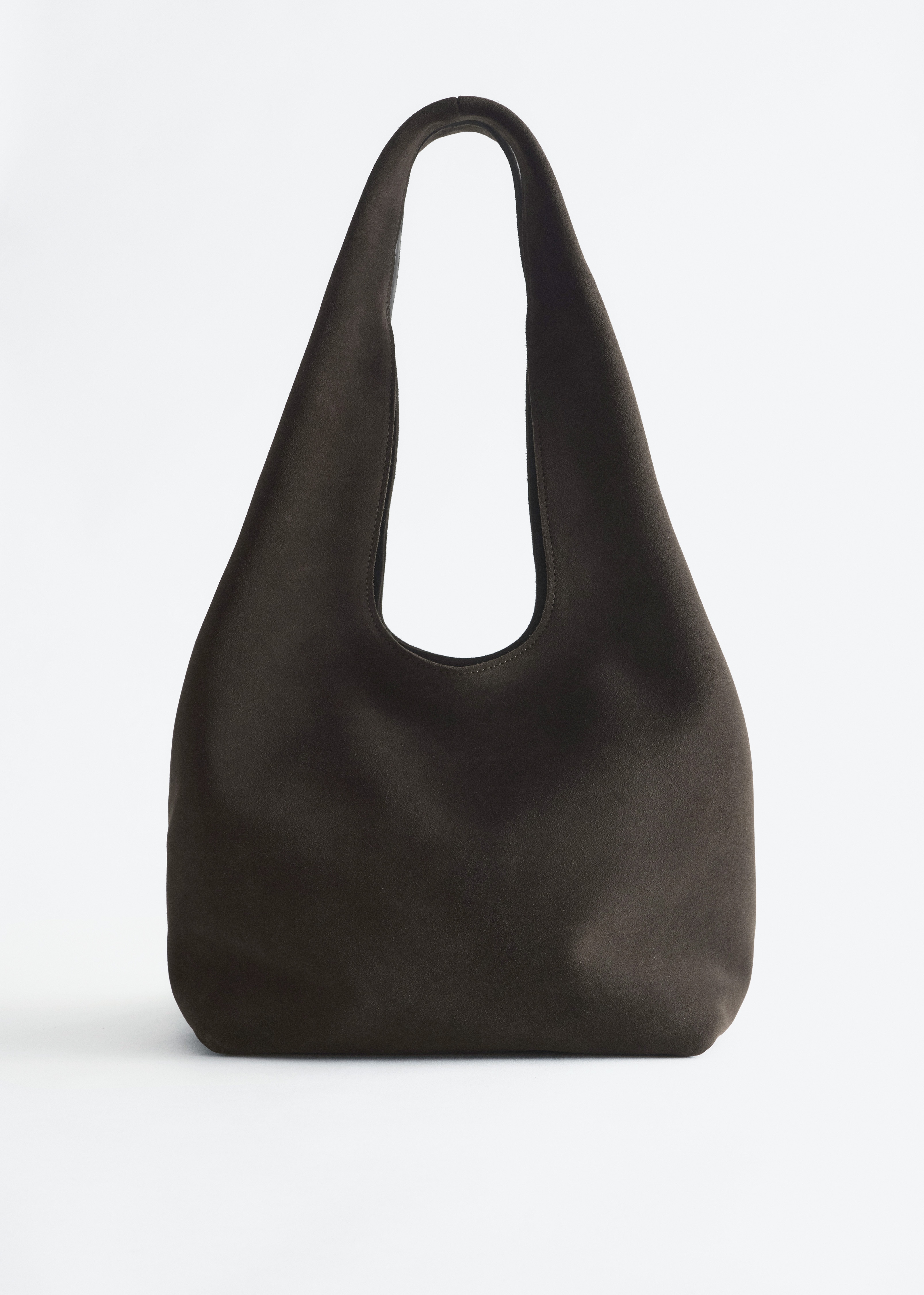 Een donkergrijze totebag van gespleten suède met een ruime, nonchalante silhouette wordt over de schouder gedragen door het model, dat de brede schouderriem vasthoudt. De tas wordt gestyled met een donkergroen satijnen hemd met lange mouwen en een blauwe jeans.