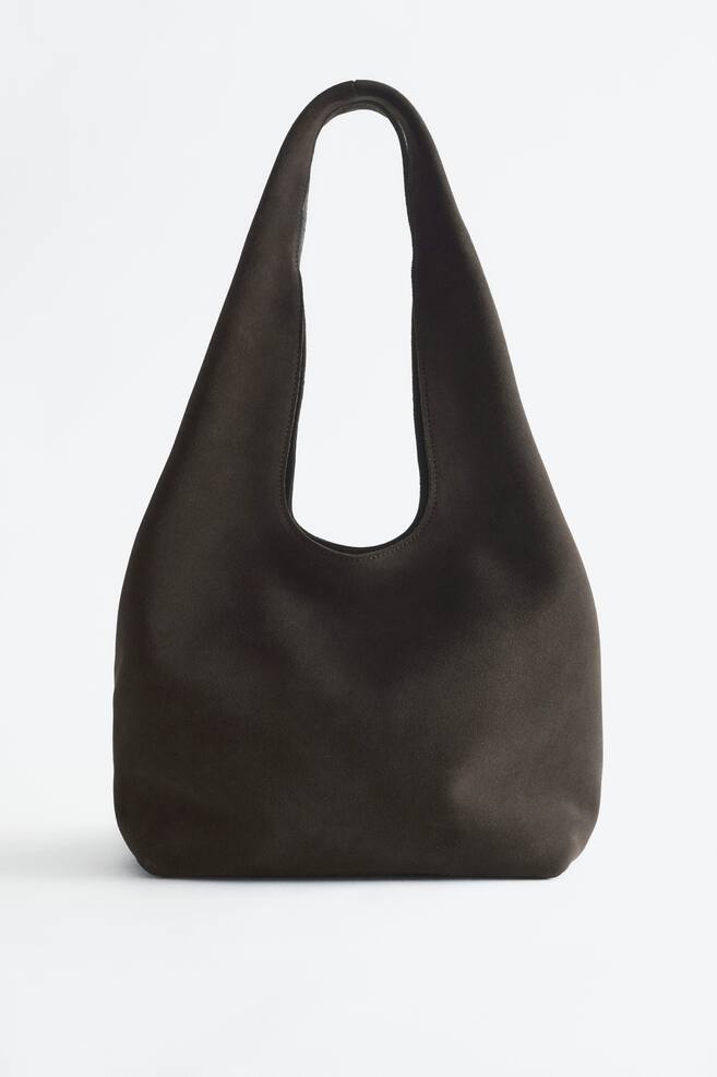 Un sac cabas en cuir suÃ©dÃ© fendu marron foncÃ© est prÃ©sentÃ© de face, mettant en valeur sa silhouette arrondie et souple ainsi que sa large bandouliÃ¨re intÃ©grÃ©e. Le sac arbore un design minimaliste et Ã©purÃ©, avec une texture mate douce et aucune quincaillerie visible. - 1