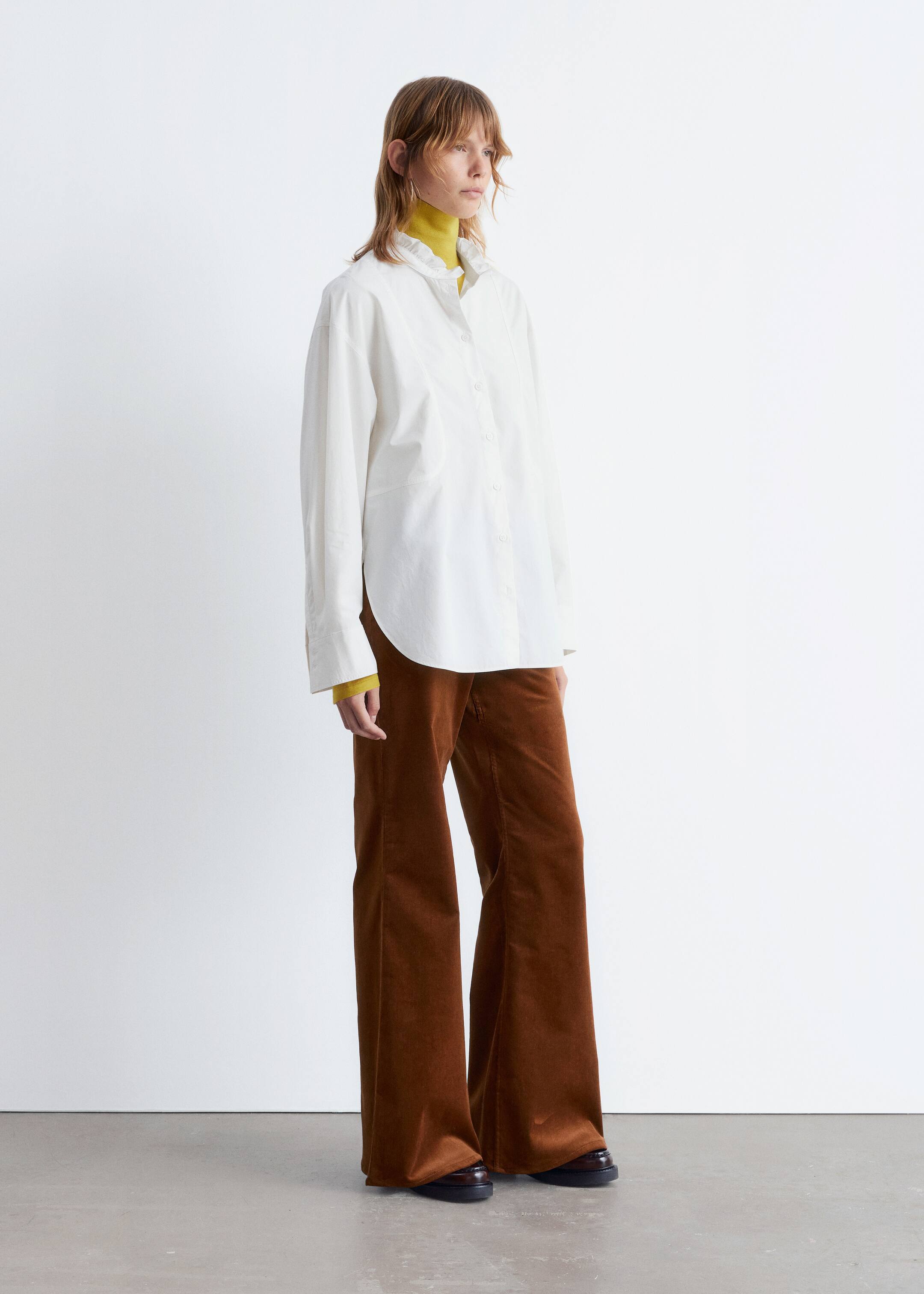 Ver imagen más grande: Una camisa blanca de gran tamaño con botones muestra un cuello con volantes de pie y puños anchos, con un dobladillo redondeado visible en el lateral. Se lleva sobre un top amarillo de cuello alto, combinado con pantalones de pana marrones de pierna ancha que cubren zapatos oscuros.