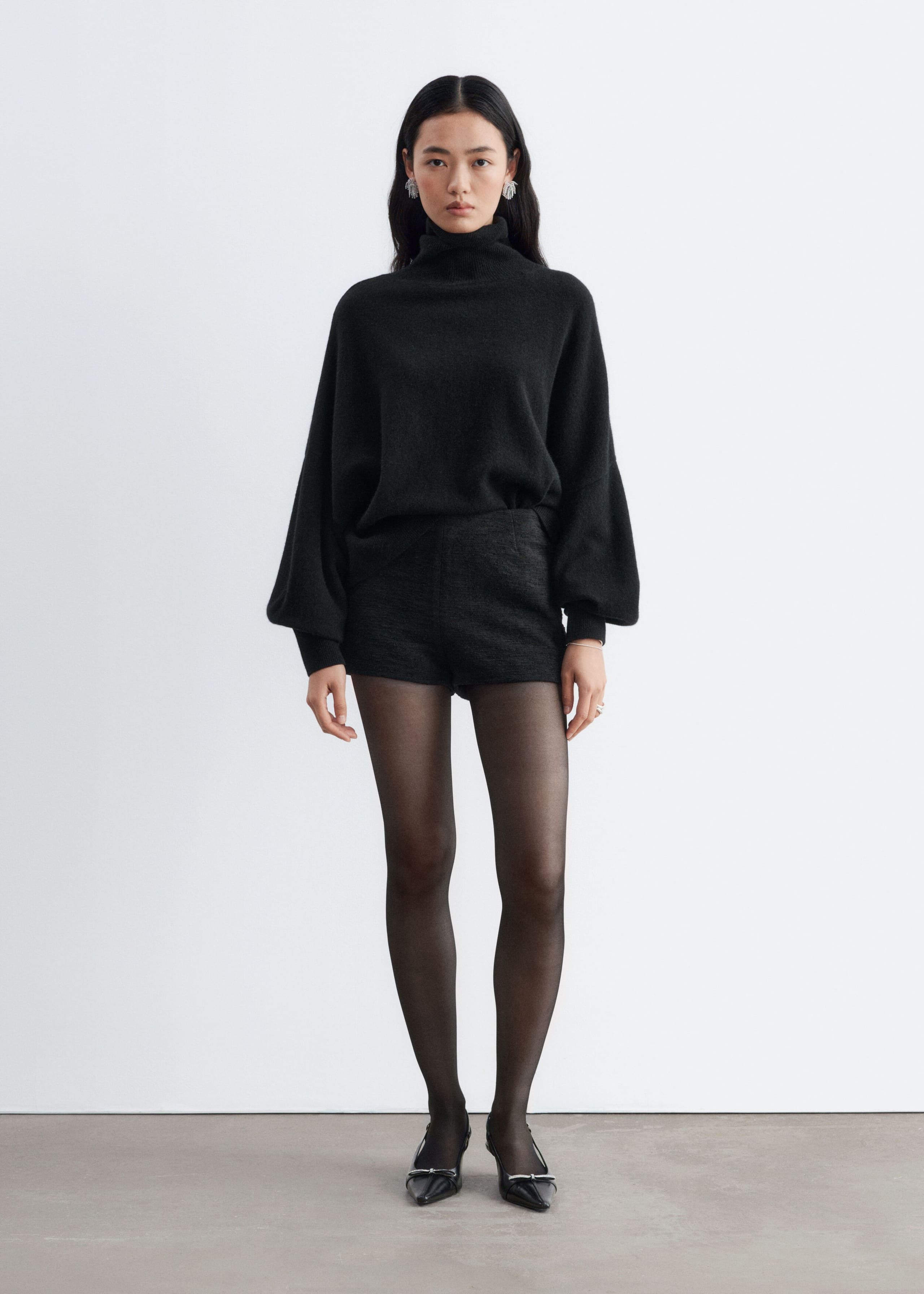 Dark grey tweed shorts with a structured, high-waisted fit are being worn by the model. The shorts are styled with a black roll-neck sweater partially tucked into the waistband, sheer black tights, and black pointed-toe flats.