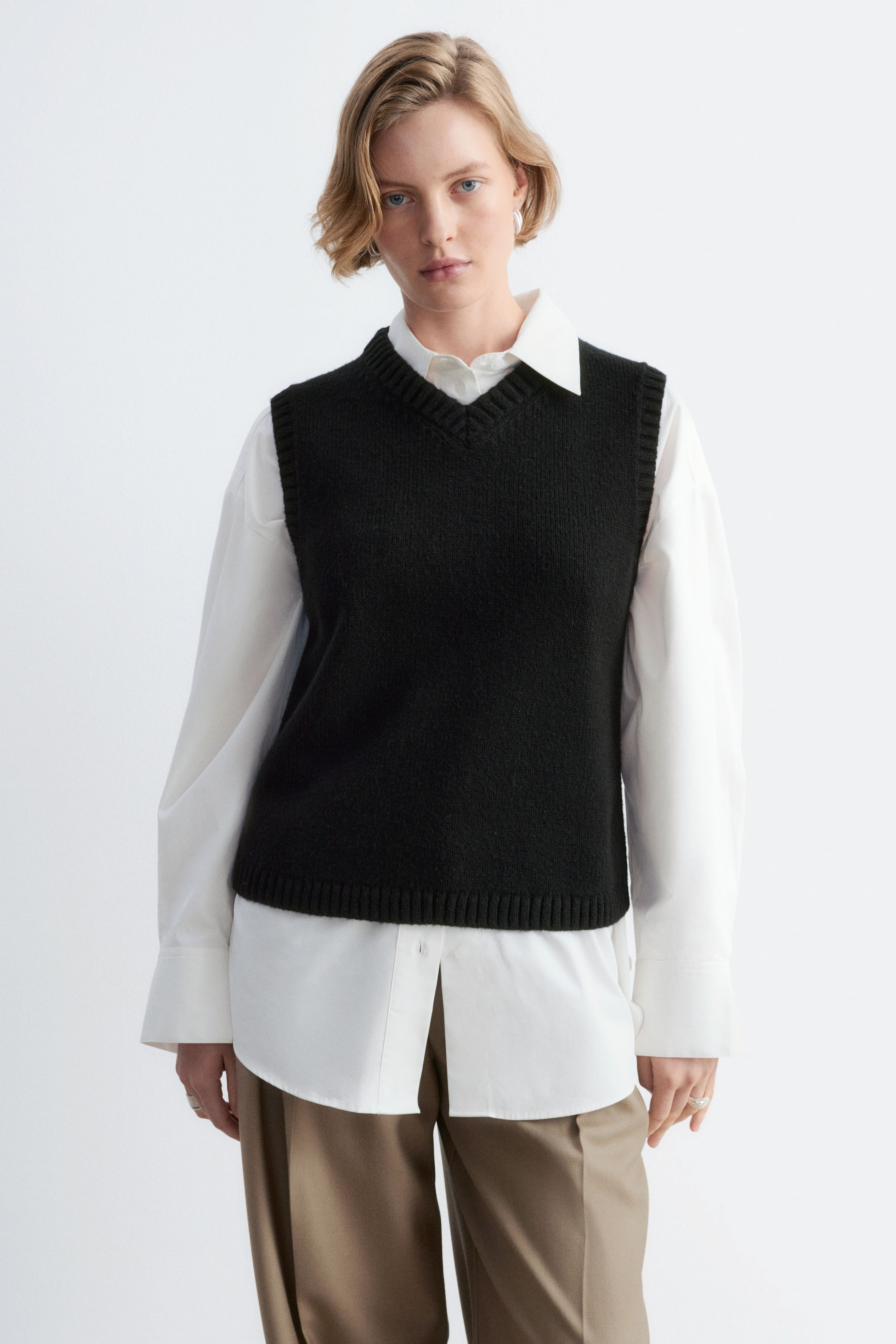 Agrandir l'image: Un gilet en maille noir en laine et coton, doté d’un col en V et de finitions côtelées, est porté par le mannequin. Le gilet est superposé à une chemise blanche à manches longues avec col, et associé à un pantalon beige.