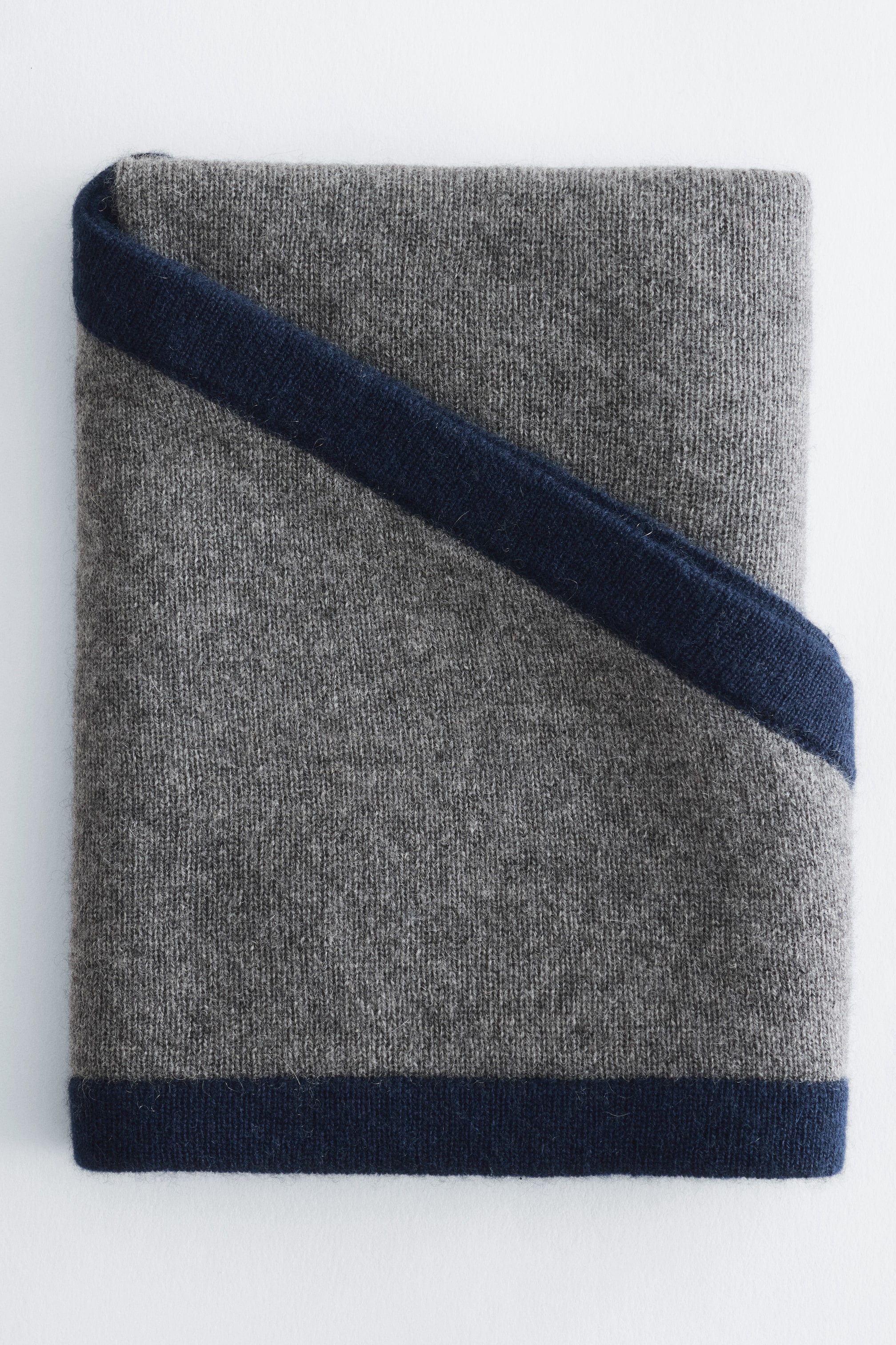 Grösseres Bild ansehen: Ein Dreieckstuch ist zu einer rechteckigen Form gefaltet und zeigt einen mittelgrauen Strickstoff. Dunkelblaue kontrastierende Ränder sind entlang der unteren Kante und diagonal über die obere rechte Falte sichtbar.