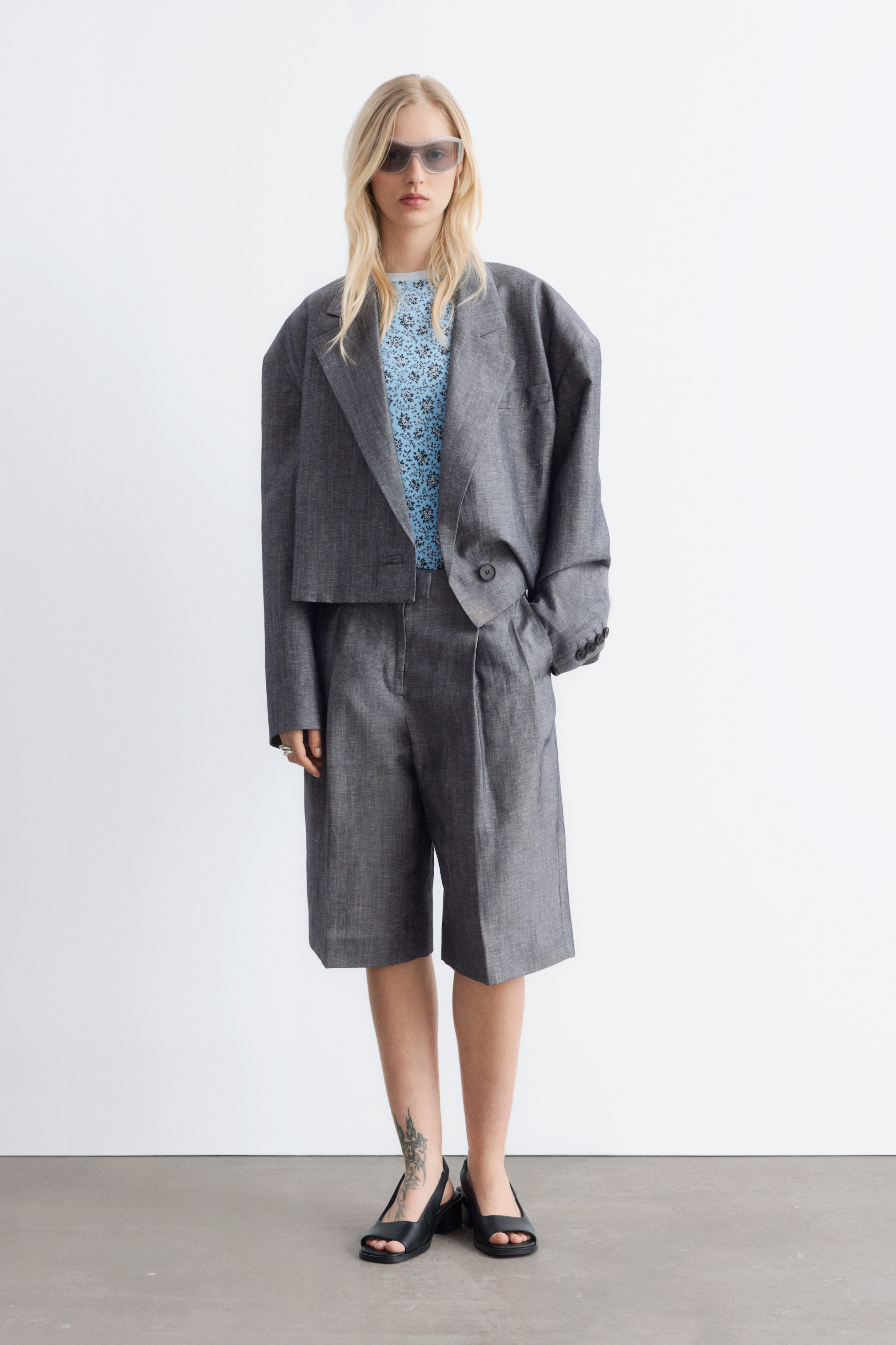 View larger image: A grey cropped blazer featuring wide notch lapels, an oversized fit with loose sleeves, and a single visible button fastening, worn open over a light blue patterned top. Matching grey knee-length shorts present a wide-leg cut with front pleats and side pockets.