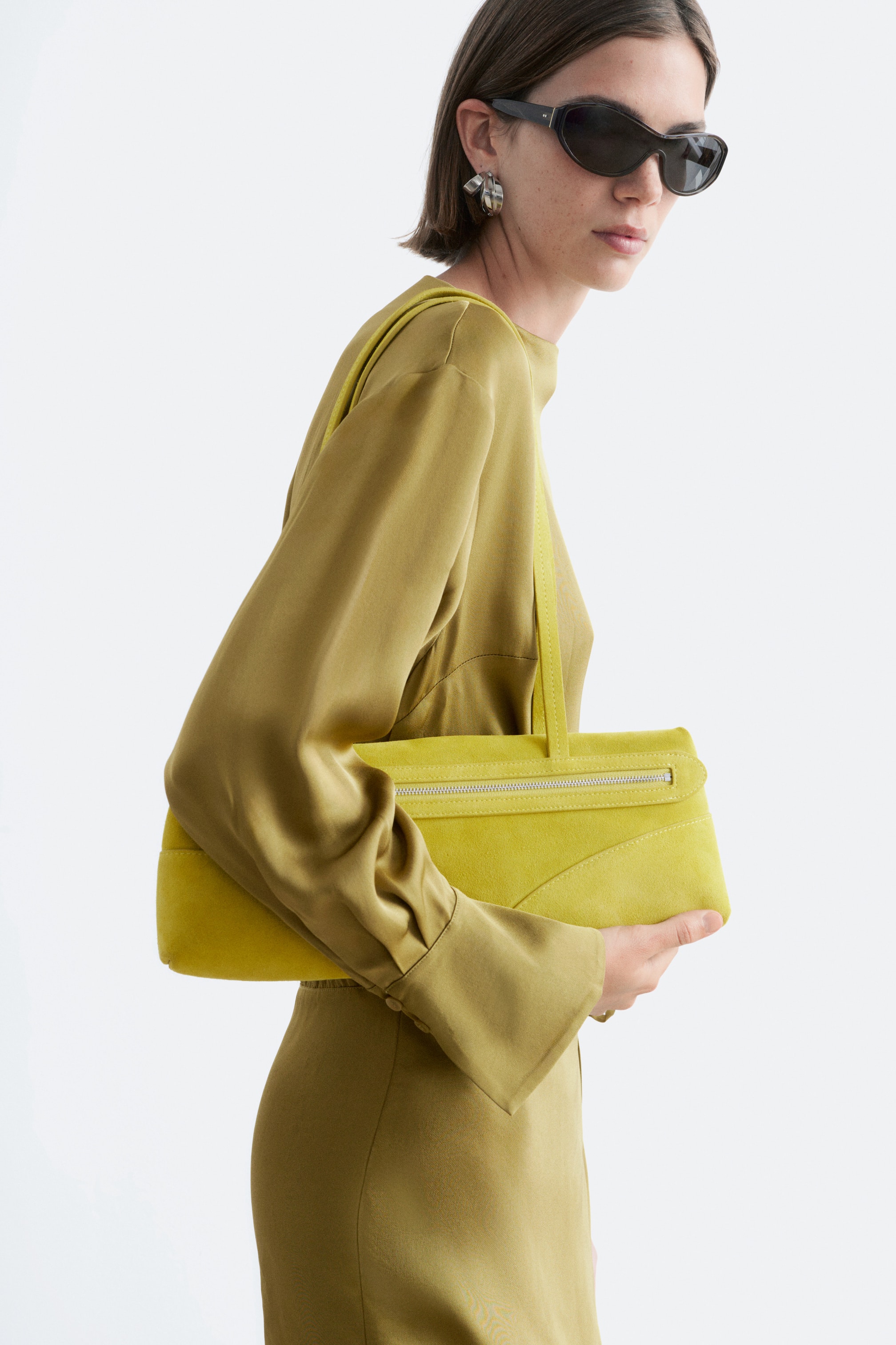 Agrandir l'image: Un sac à main rectangulaire vert citron, confectionné dans un matériau à la texture douce, est tenu sur le côté. Il présente une poche zippée au ton argenté sur son rabat avant et un détail de couture incurvée sur l'ensemble de son corps, avec une unique sangle assortie reposant sur l'épaule.