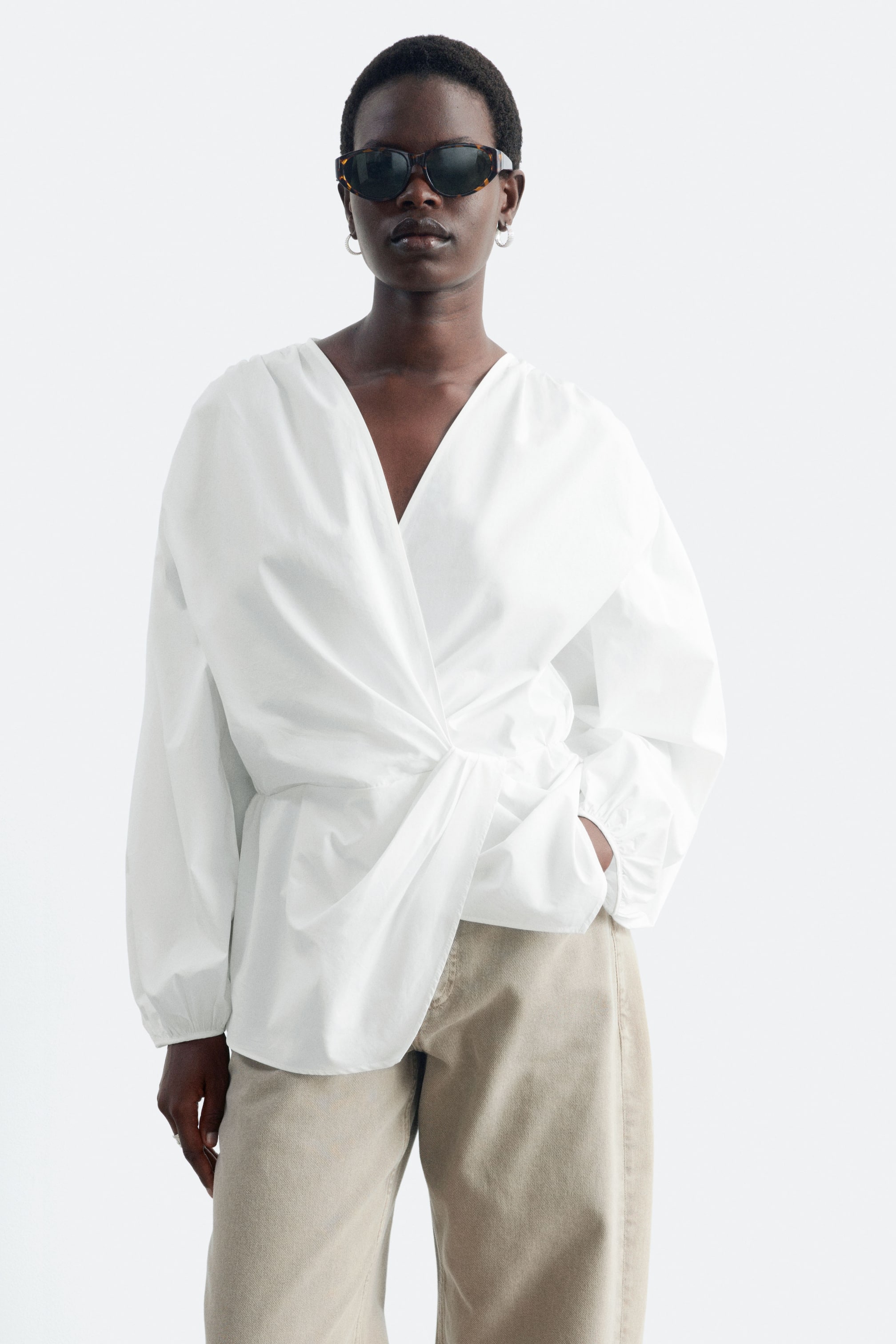 View larger image: A white cotton crossover blouse featuring a deep V-neckline, long voluminous sleeves and a gathered twist detail at the waist is worn by the model. The blouse is styled with light beige trousers, with the model resting one hand in a pocket.