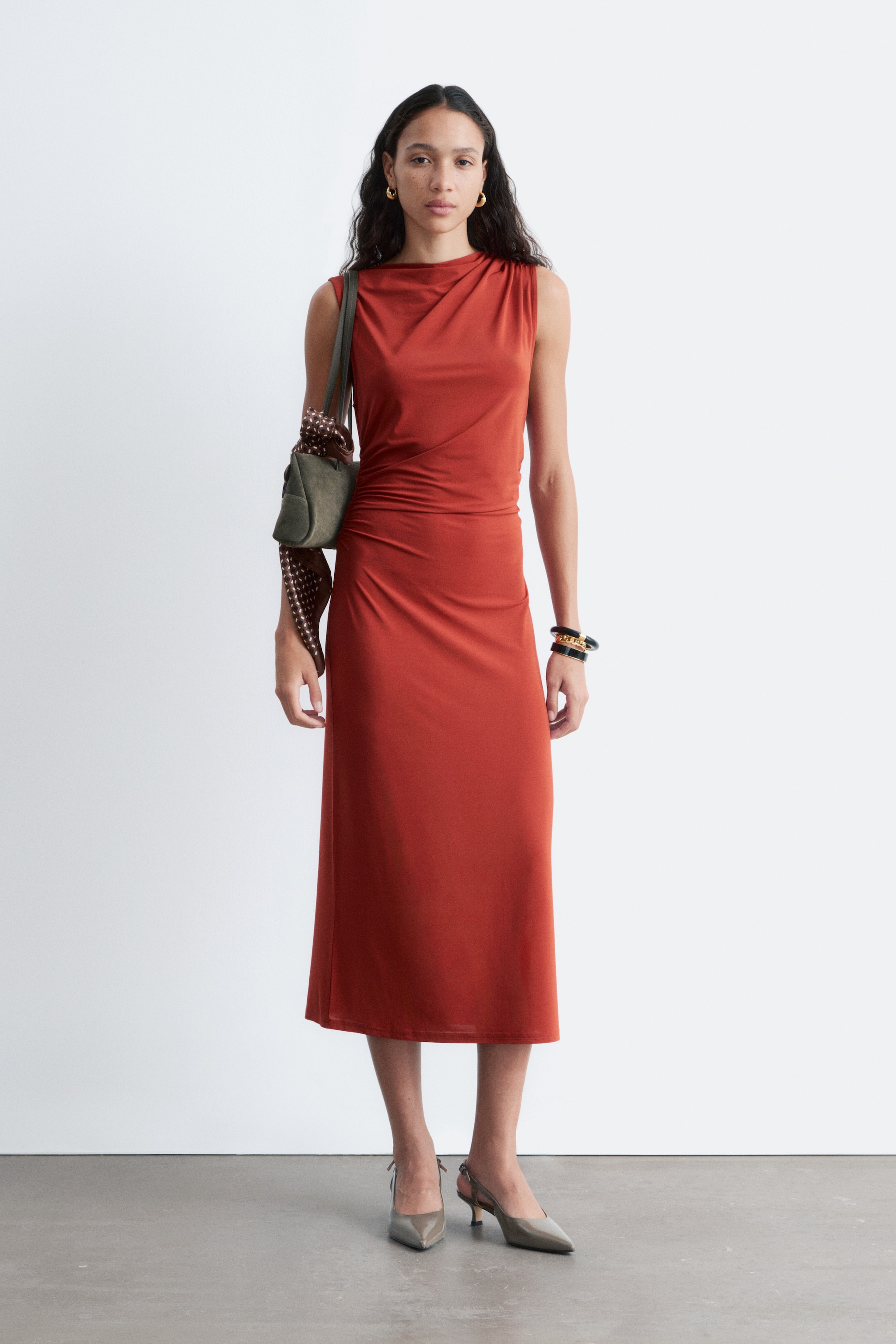 Vezi imaginea mai mare: O rochie midi fără mâneci, de culoare ruginiu, prezintă un decolteu drapat în stil barcă și adunări subtile pe partea stângă a corsajului său mulat, cu fusta curgând într-o siluetă dreaptă midi.