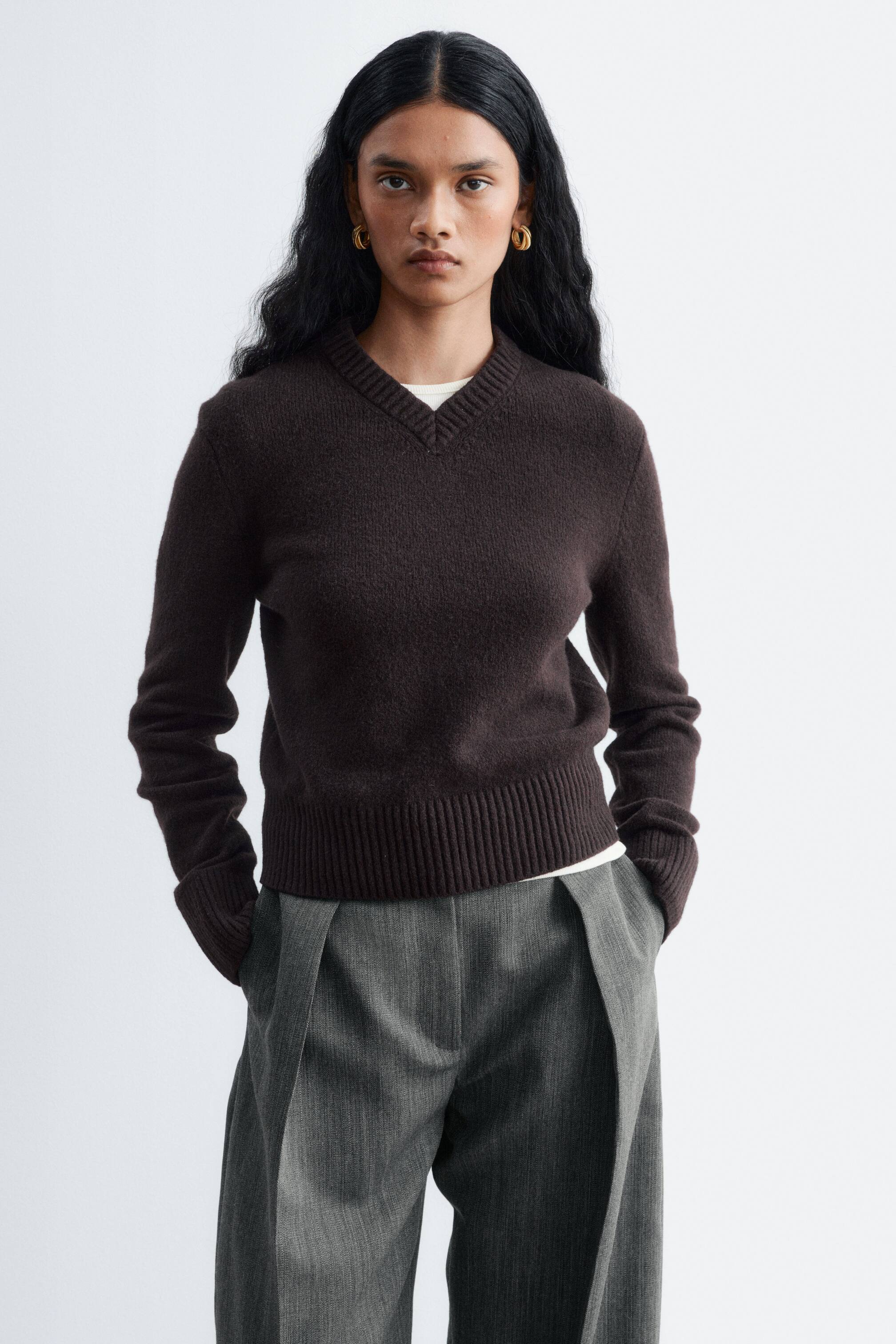 Visualizza l'immagine ingrandita: Un maglione lavorato a maglia di colore marrone scuro, con scollo a V a coste, polsini a coste e ampio orlo a coste, indossato dalla modella. Il maglione è sovrapposto a una T-shirt bianca ed è abbinato a pantaloni grigi a gamba larga.