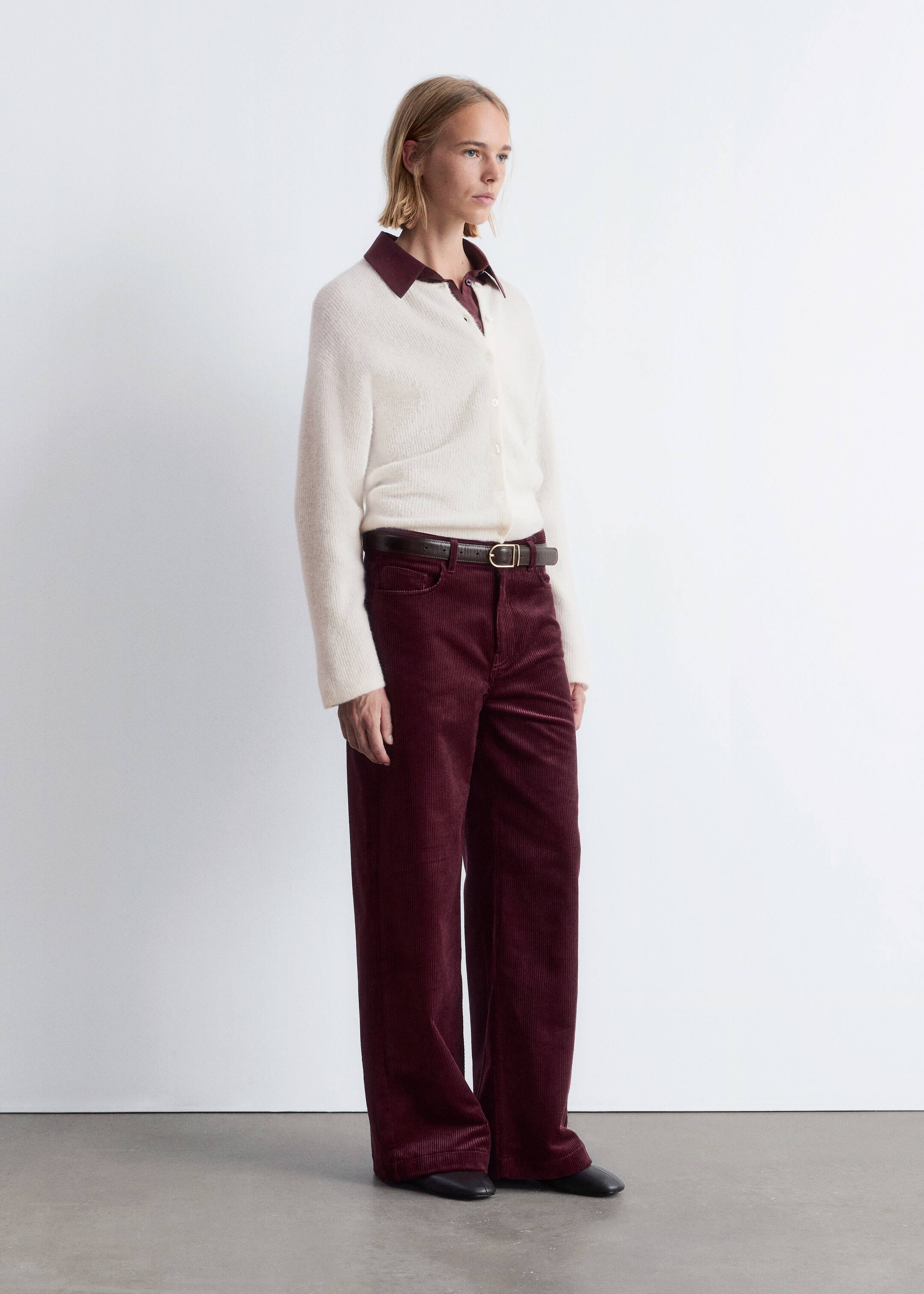 Visualizza immagine più grande: Un cardigan lavorato a maglia color bianco sporco con chiusura a bottoni è indossato sopra una camicia con colletto color bordeaux scuro, abbinato a pantaloni a gamba larga e a tutta lunghezza in velluto a coste bordeaux. Una cintura in pelle marrone scuro stringe i pantaloni in vita, e delle scarpe piatte nere a punta completano l'insieme, il tutto presentato da un'angolazione di tre quarti.