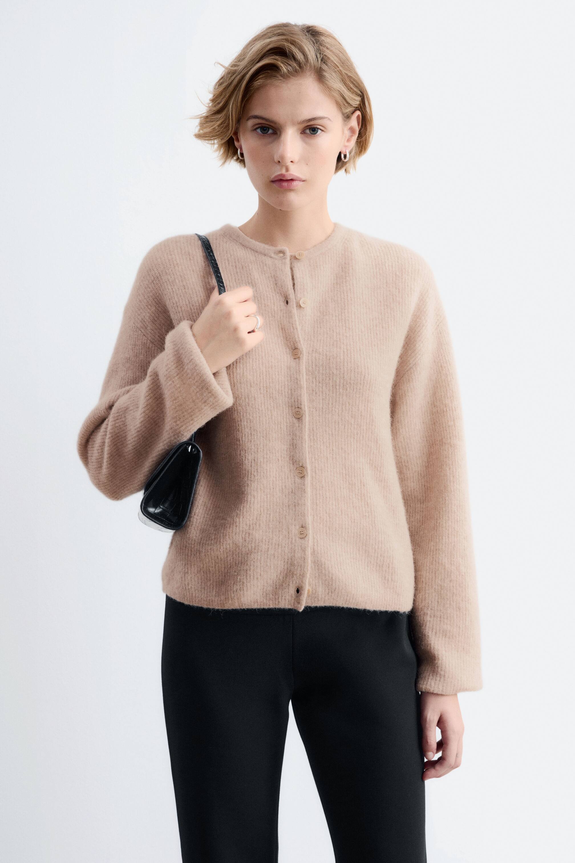 Vis større bilde: En beige, langermet strikkejakke med rund hals og frontknapper bæres av modellen. Jakken er stylet med svarte bukser, og modellen holder ремmen til en svart skulderveske.